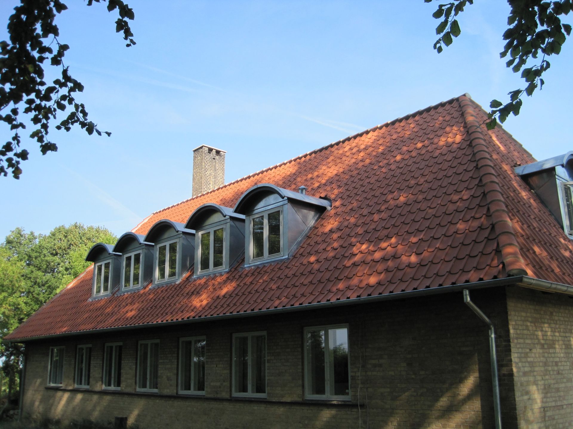 Rødt tegltag med kvistvinduer på en murstensbygning under en blå himmel, træer i baggrunden.