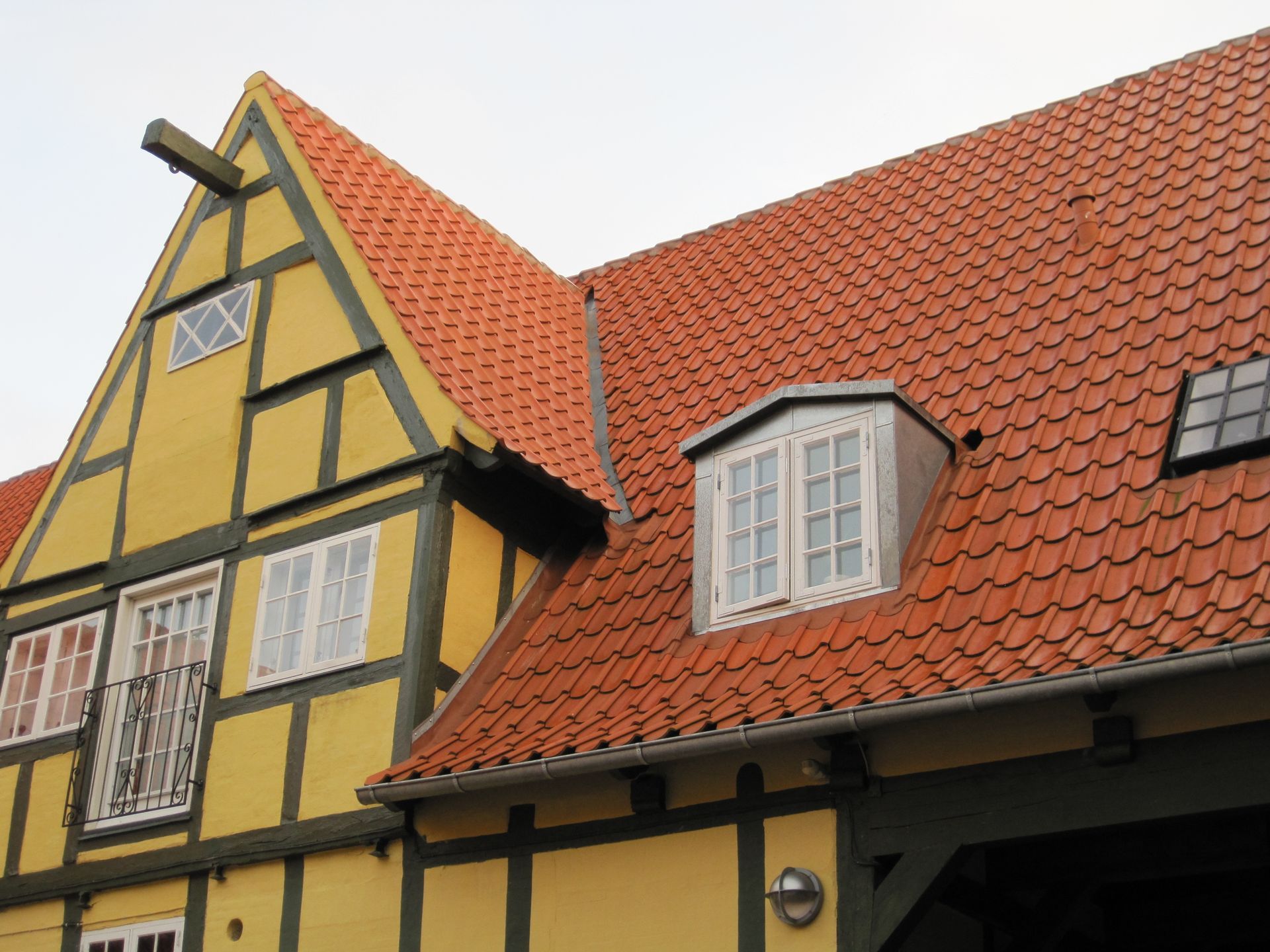 Gul og brun bindingsværksbygning med rødt tegltag, kvistvindue og lille balkon.