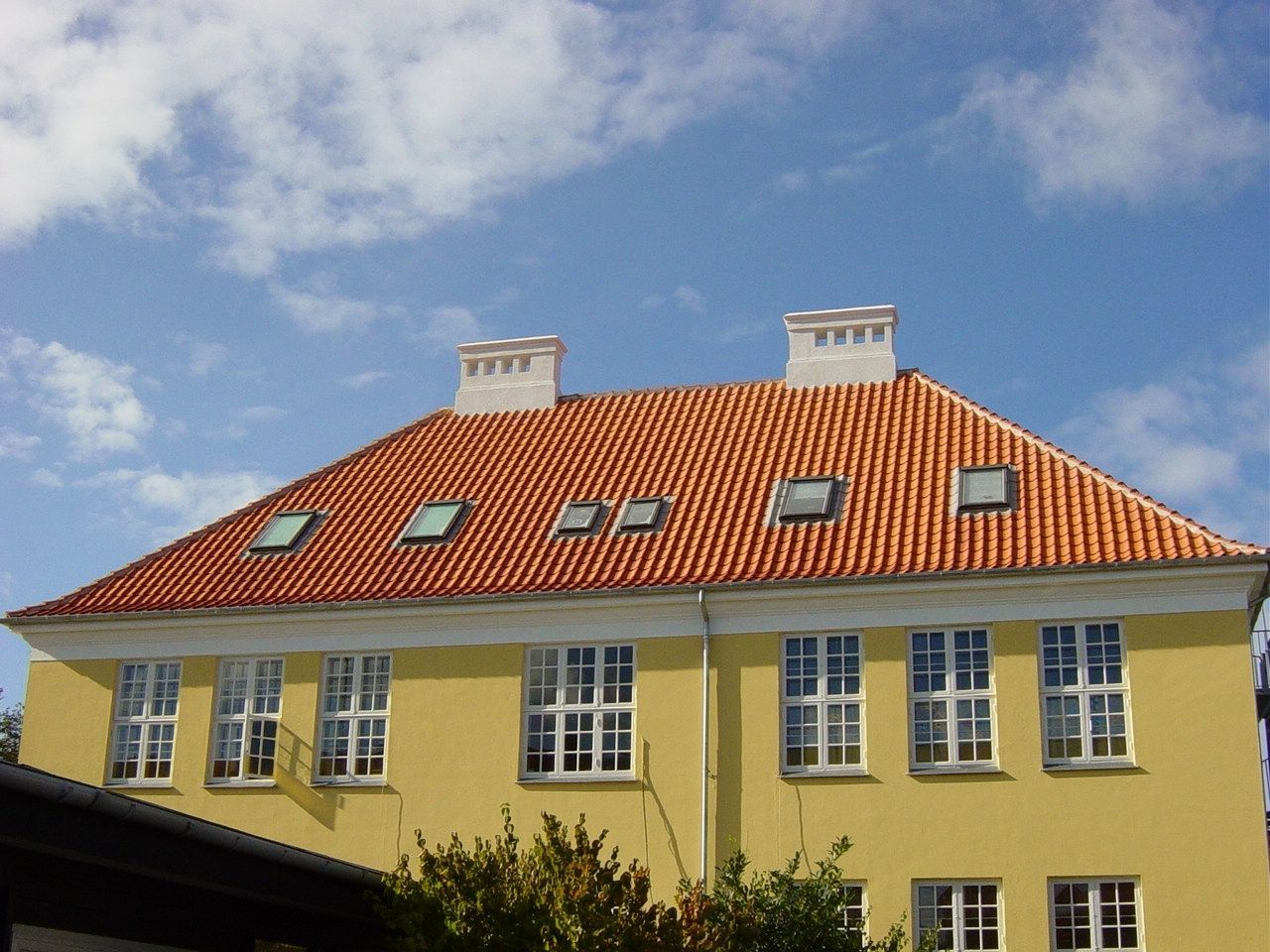Gul bygning med orange tegltag, flere vinduer og ovenlysvinduer mod en blå himmel.