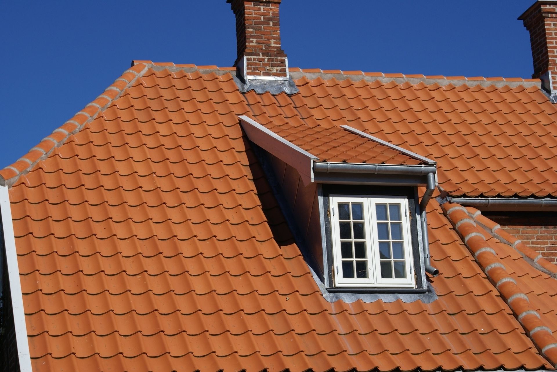 Rødt tegltag med kvistvindue, skorstene og blå himmel i baggrunden.
