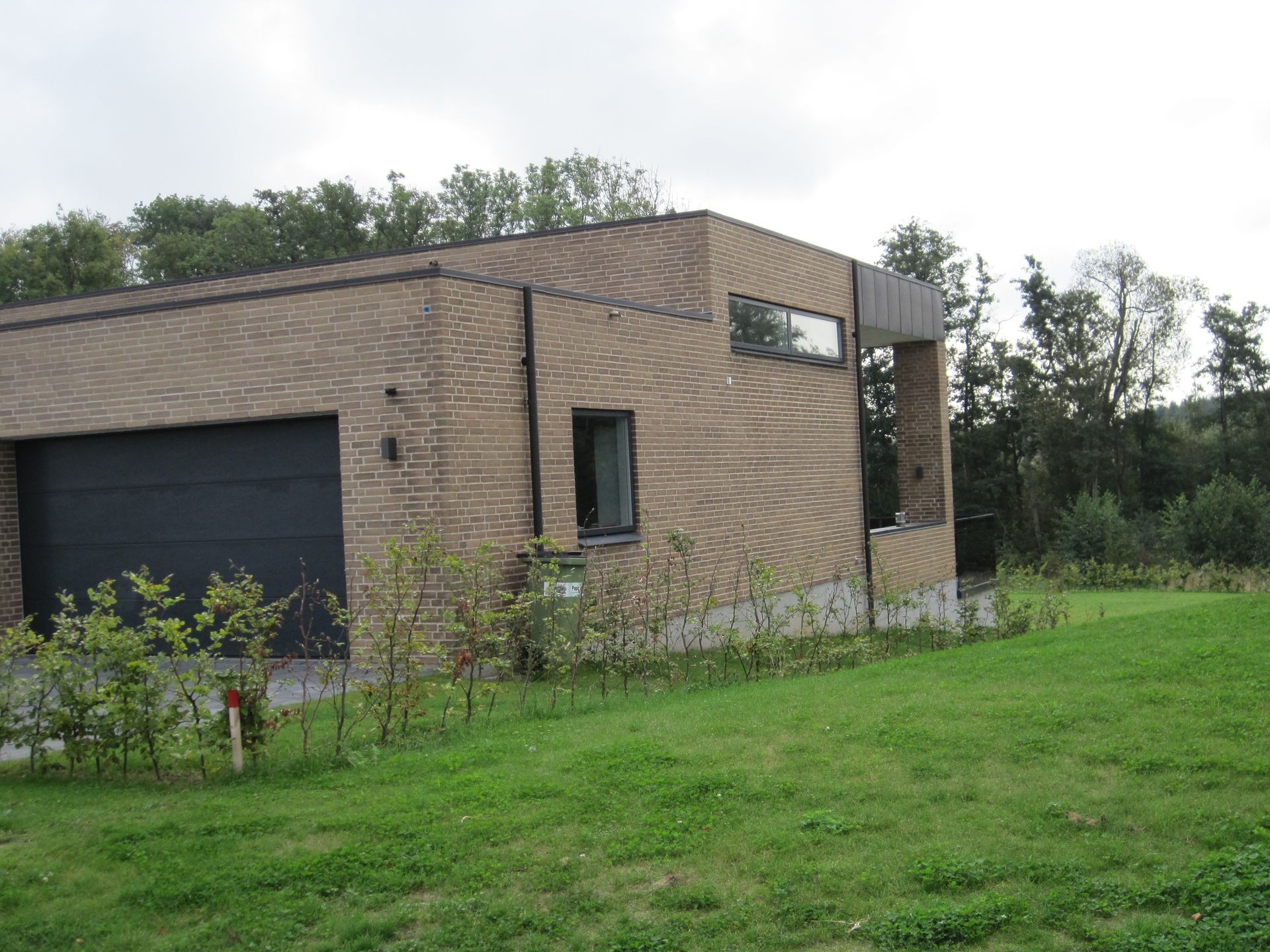 Brun murstensbygning med en sort garageport og to vinduer, beliggende på en grøn græsplæne med træer i baggrunden.