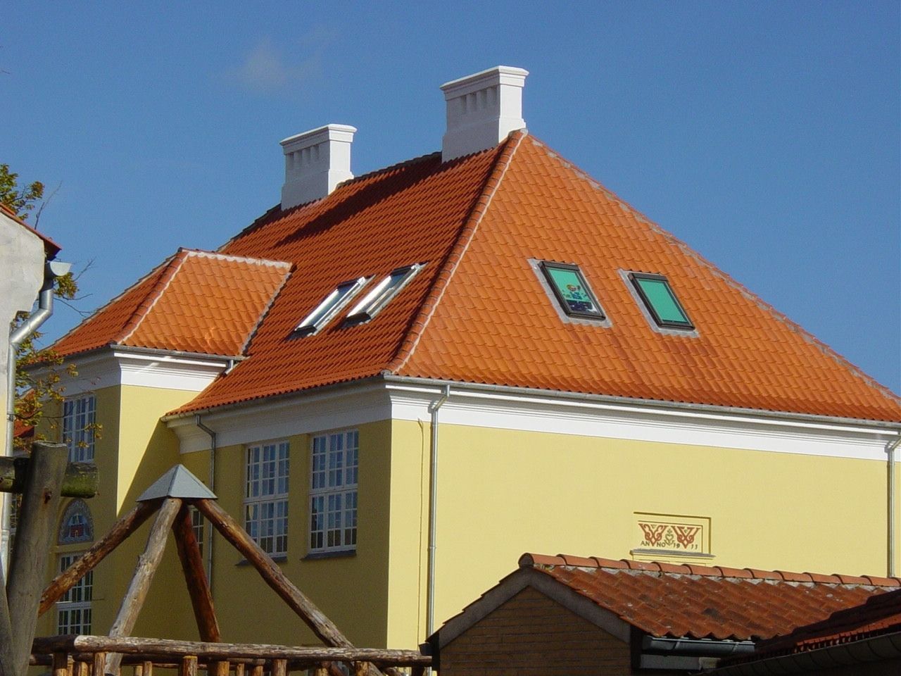 Gul bygning med rødt tegltag, hvide lister, skorstene og ovenlysvinduer mod en blå himmel.
