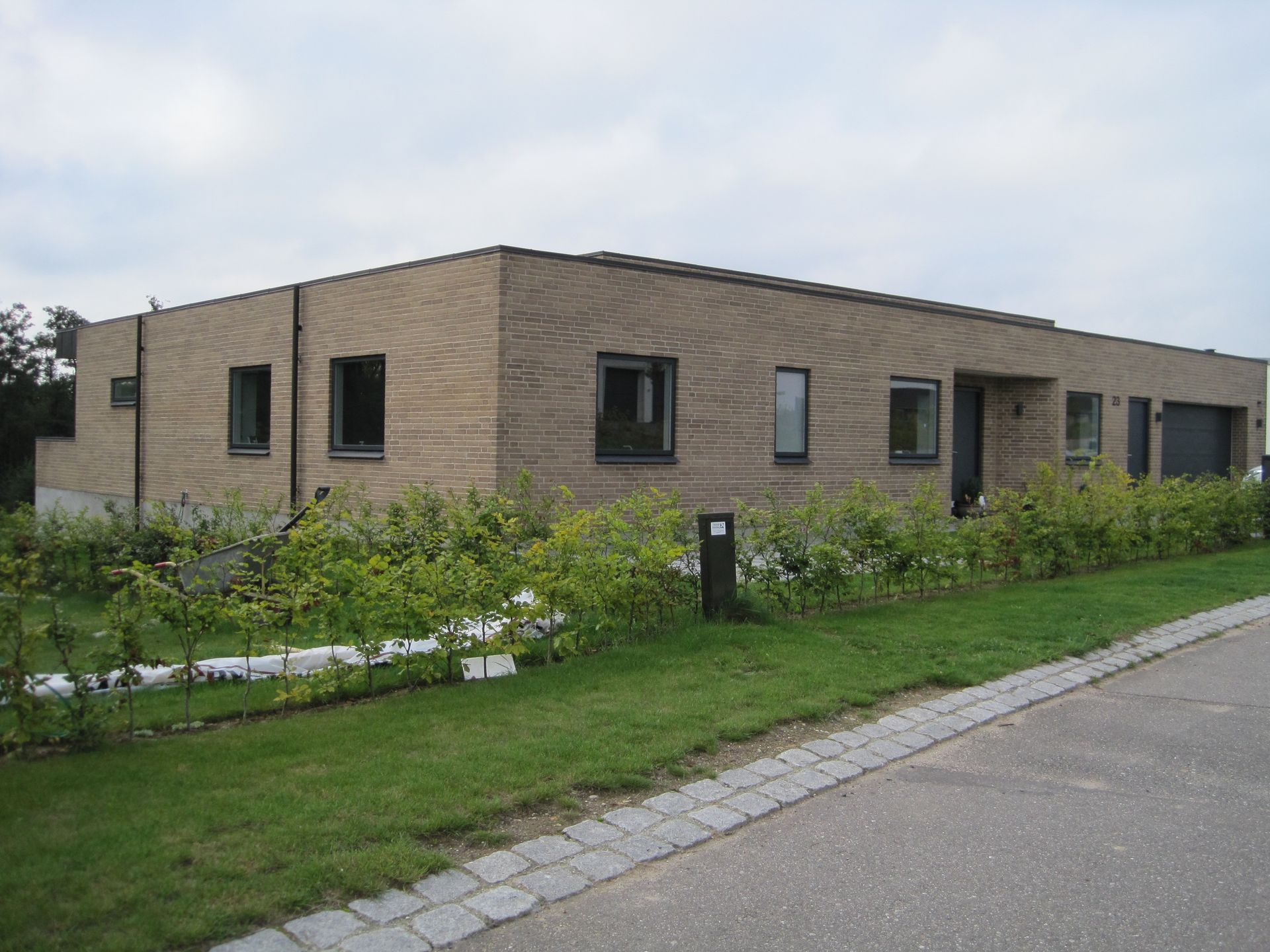 Moderne etplans murstenshus med mørke vinduer og en lille forhave med buske. Overskyet himmel.