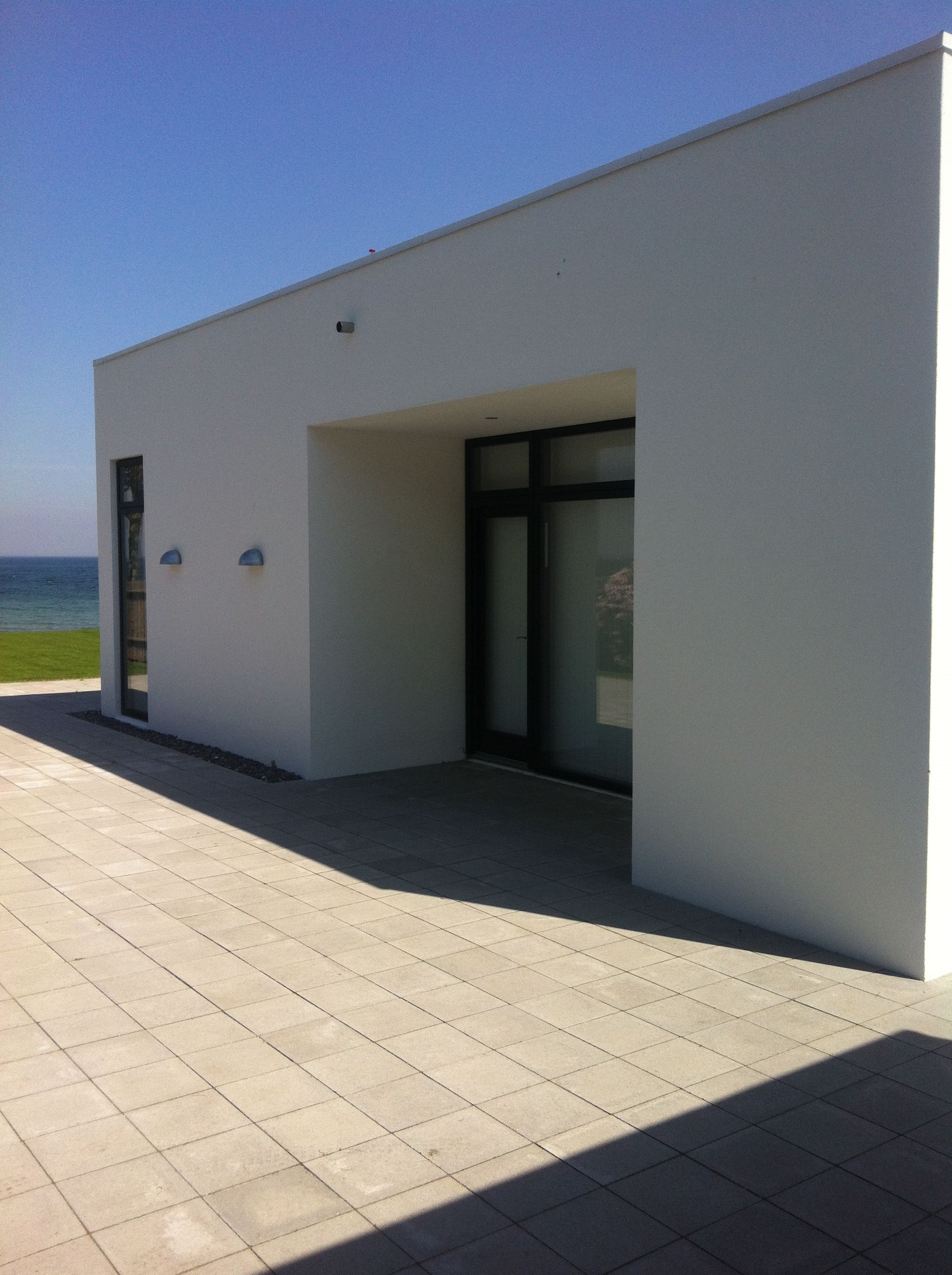 Hvid bygning med mørkrammet dør og vinduer. Flisebelagt terrasse med havudsigt under en klar blå himmel.
