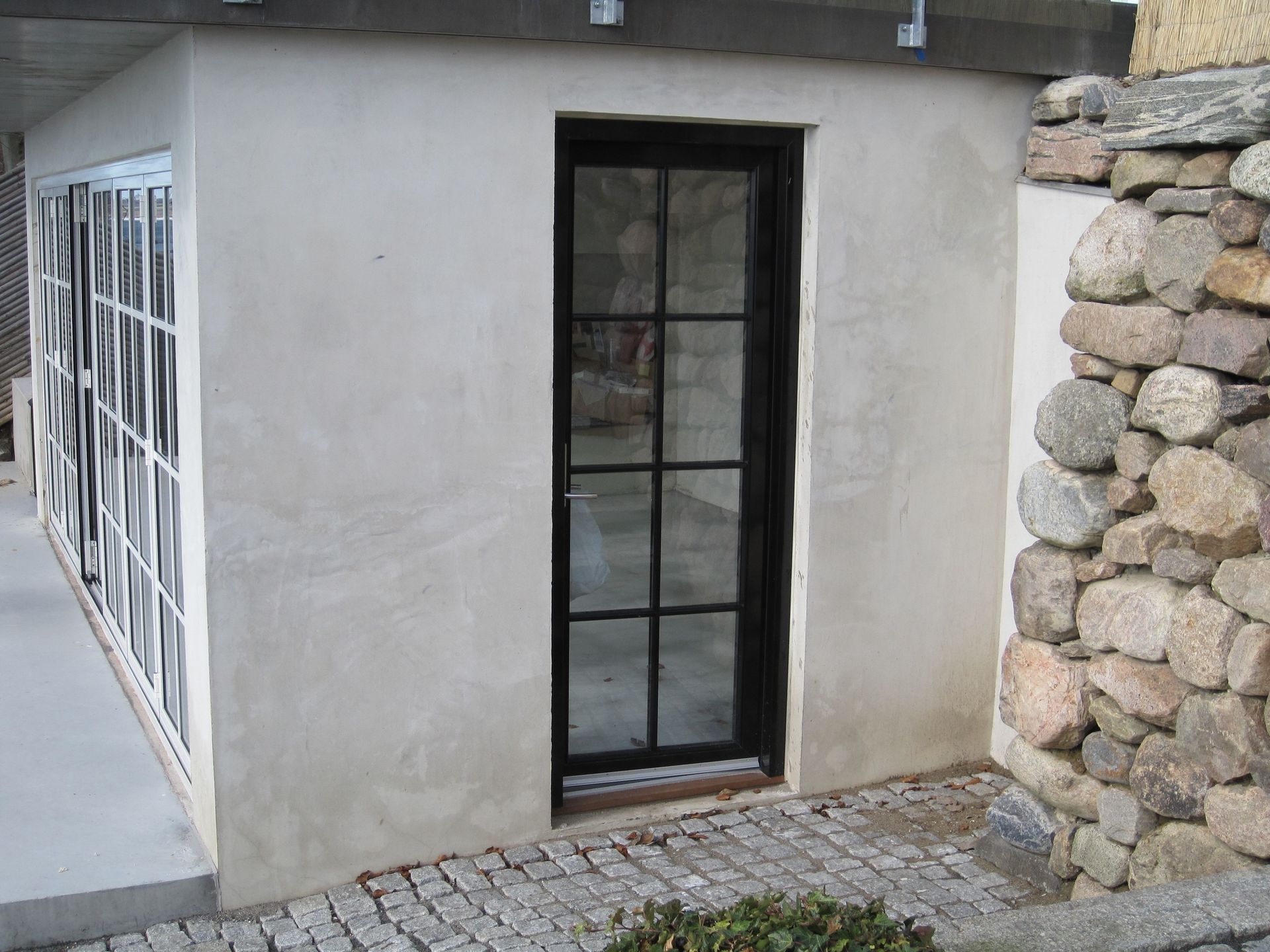 Udvendig udsigt over en bygning med en sortindrammet glasdør, en stenmur og en brostensbelagt sti.