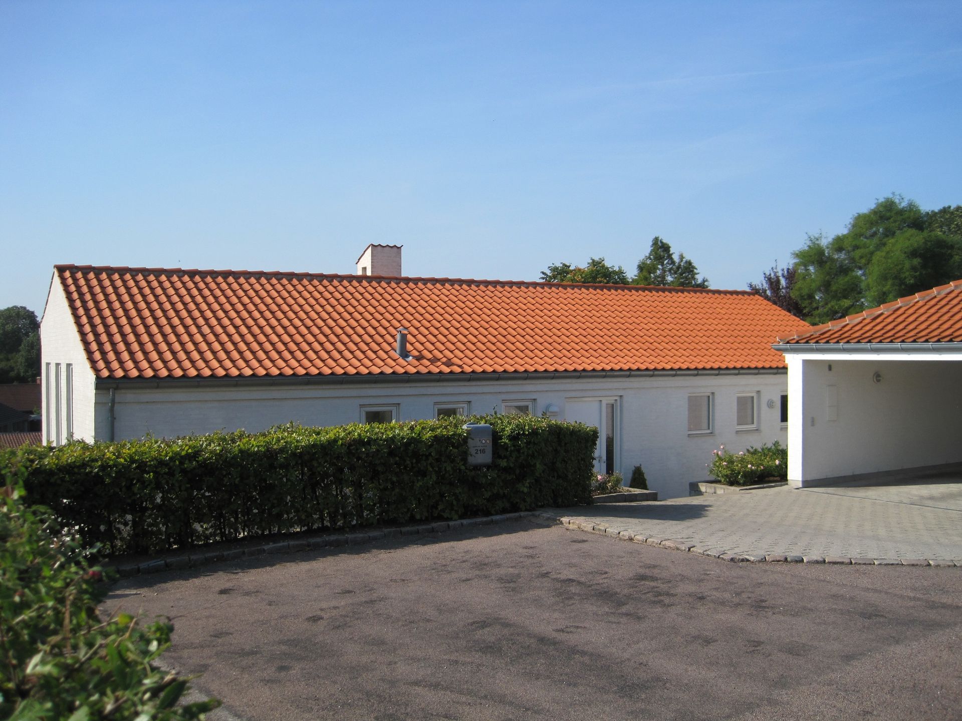 Hvidt hus med orange tegltag, grøn hæk, indkørsel og carport på en solskinsdag.