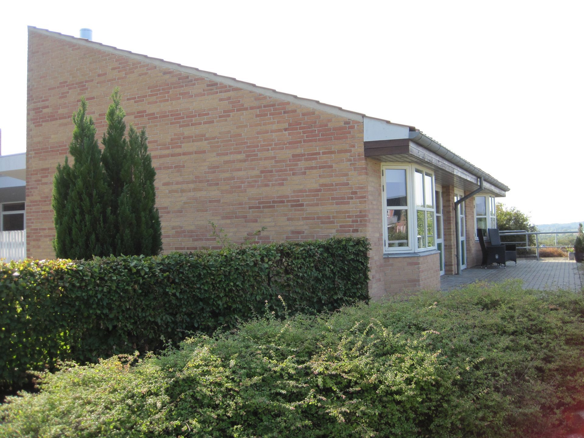 Murstensbygning med hæk og vinduer. Grønne buske i forgrunden.