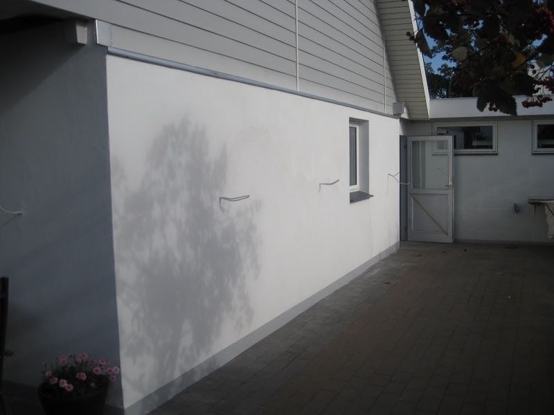 Hvid bygningsfacade med vindue og døråbning; gråt brolagt område; lyserøde blomster i potte.