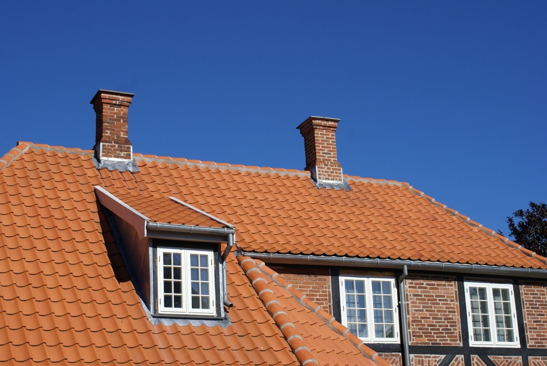 Rødt tegltag med to murstensskorstene og kvistvindue mod en klar blå himmel.