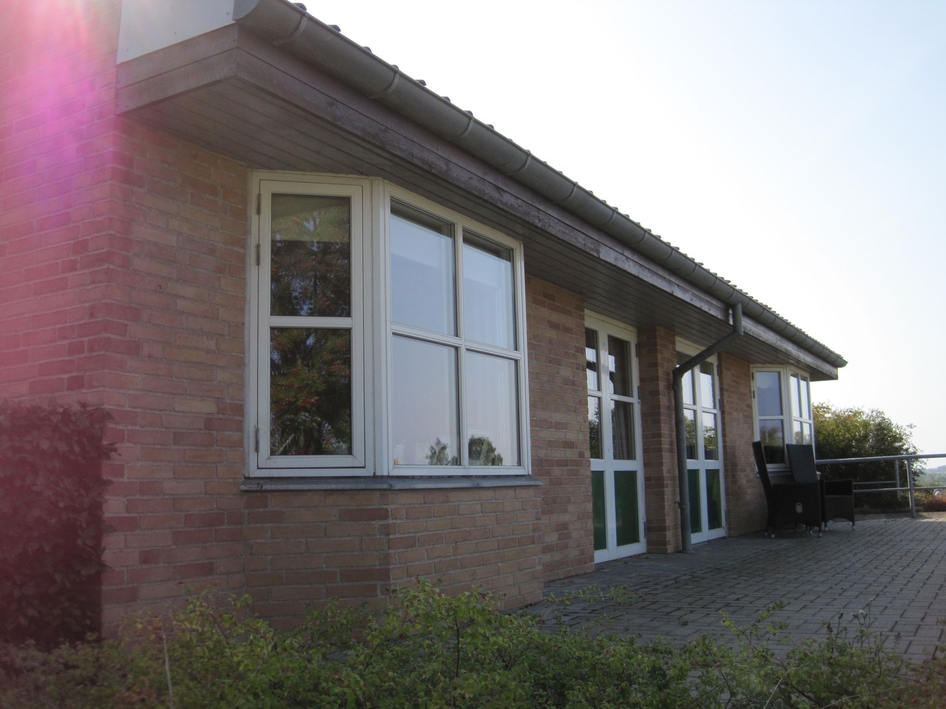 Murstensbygning med hvide indrammede vinduer, et fladt tag og en lille terrasse med grønne områder foran.