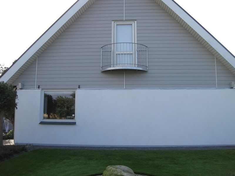 Facade af et hus med gråt tag og facadebeklædning, en lille altan og et vindue på en hvid væg.