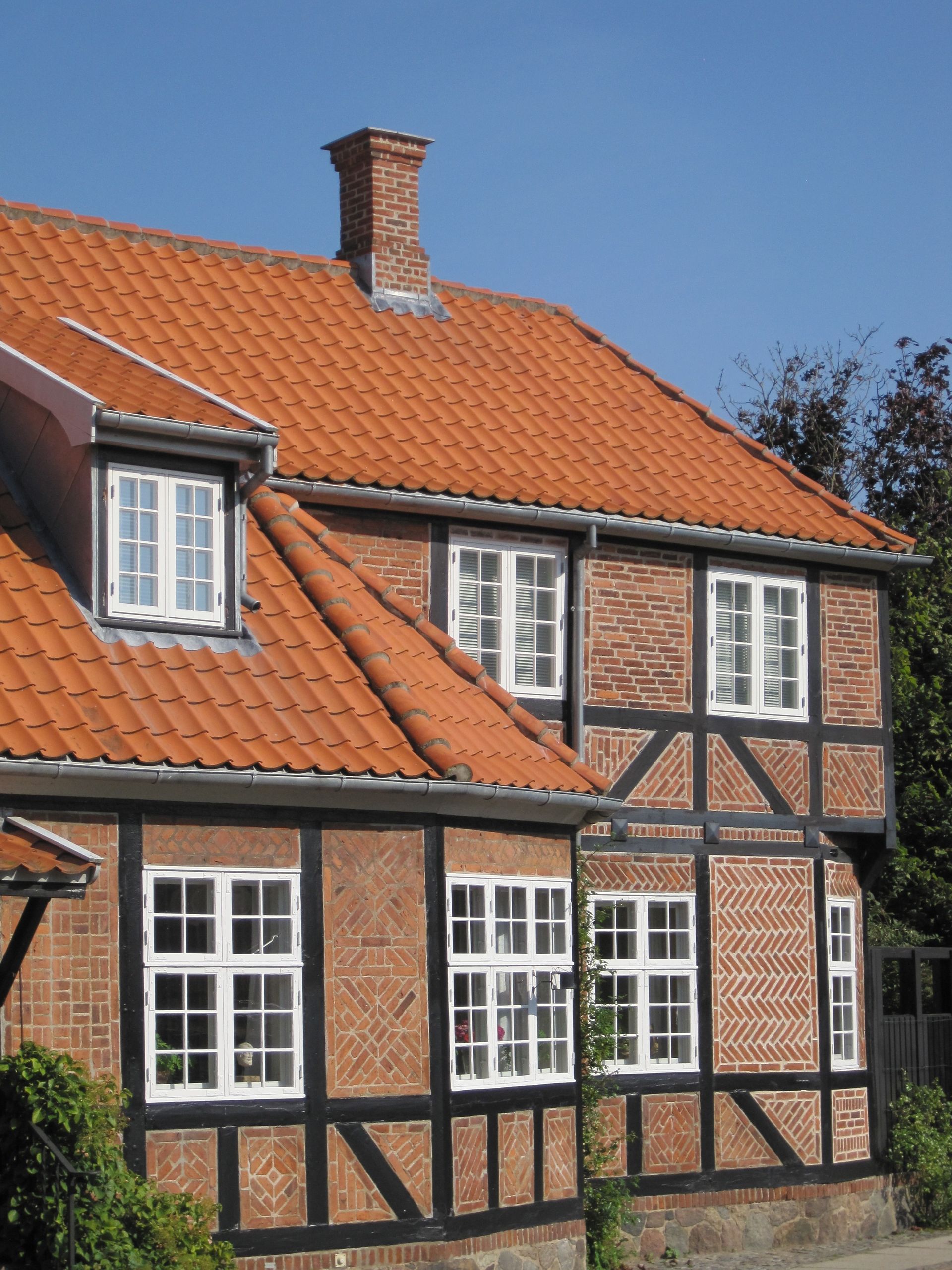 To-etagers murstensbygning med rødt tegltag, sort bindingsværkskonstruktion og hvidindrammede vinduer mod en blå himmel.