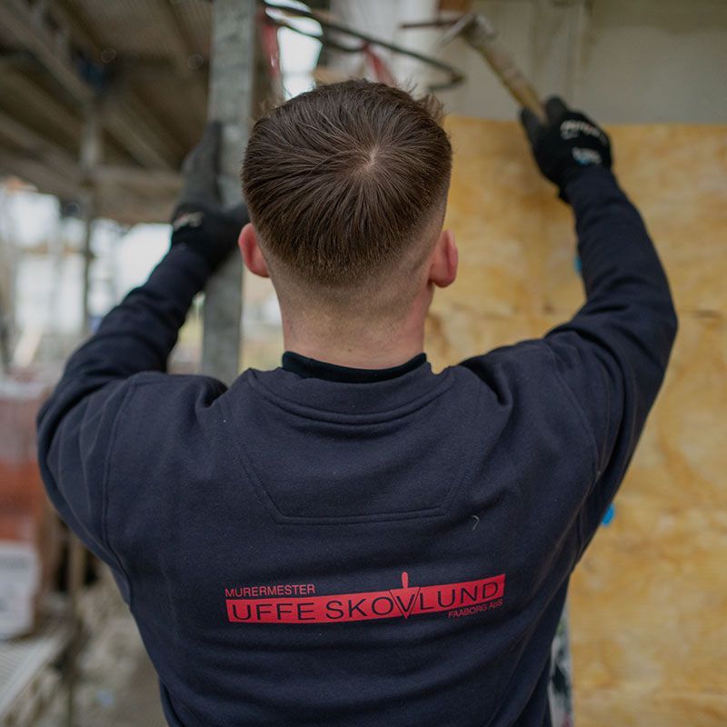 Bygningsarbejder hamrer træ, set bagfra. Iført mørkeblå skjorte og sorte handsker. Stillads i baggrunden.