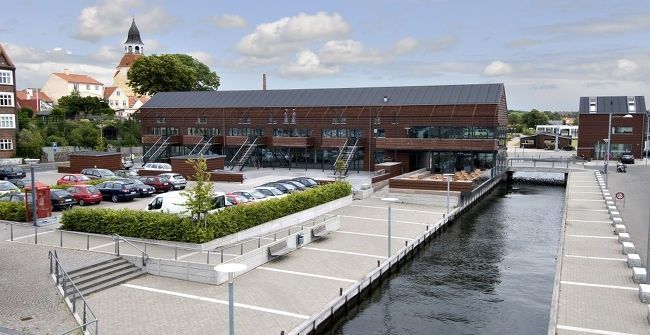 Bygning med mørkt tag og glasvinduer, promenade langs vandet med parkerede biler og en kirke i baggrunden.
