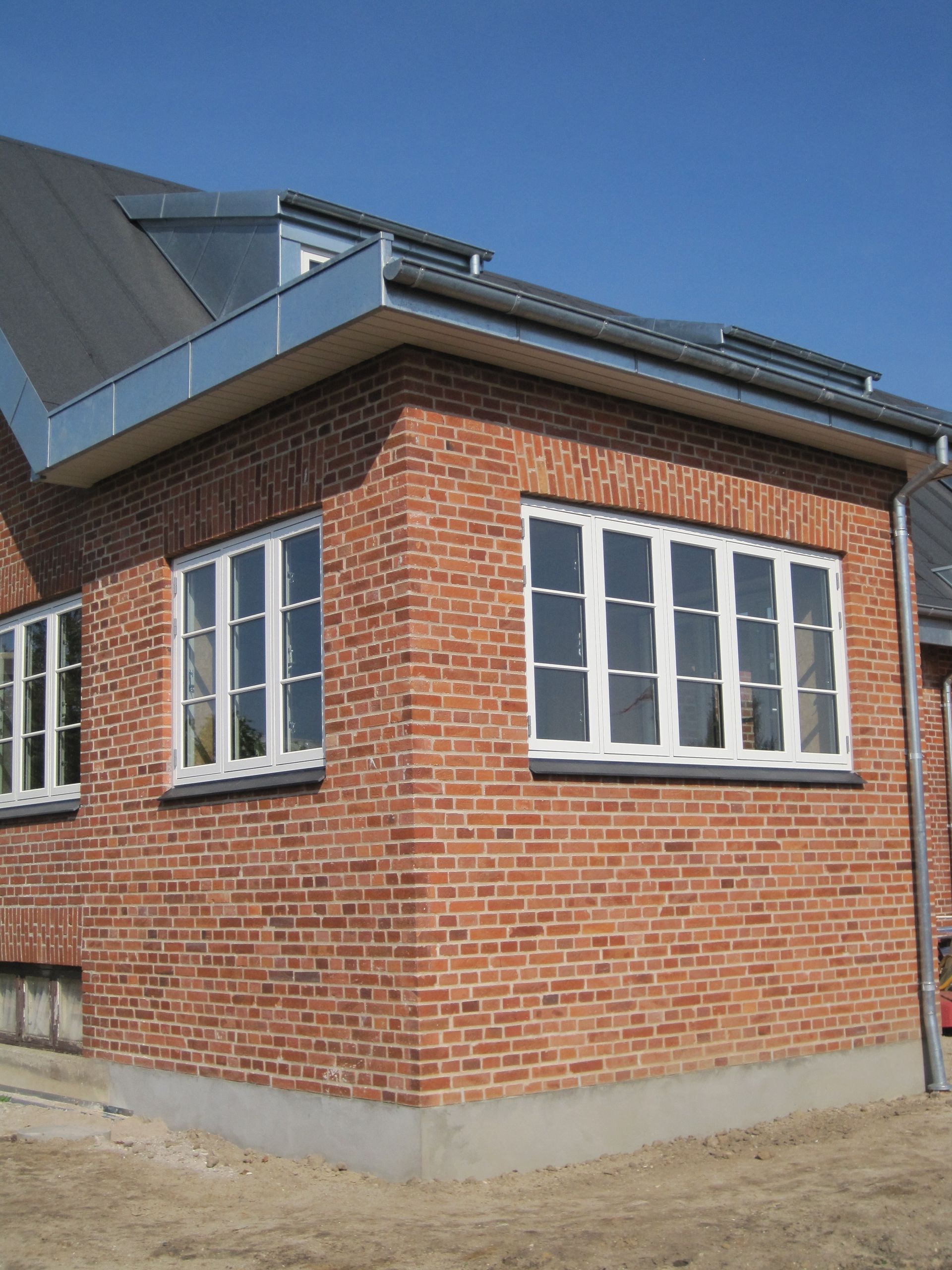 Hjørne af murstensbygning med hvidindrammede vinduer, gråt metaltag og en klar blå himmel.