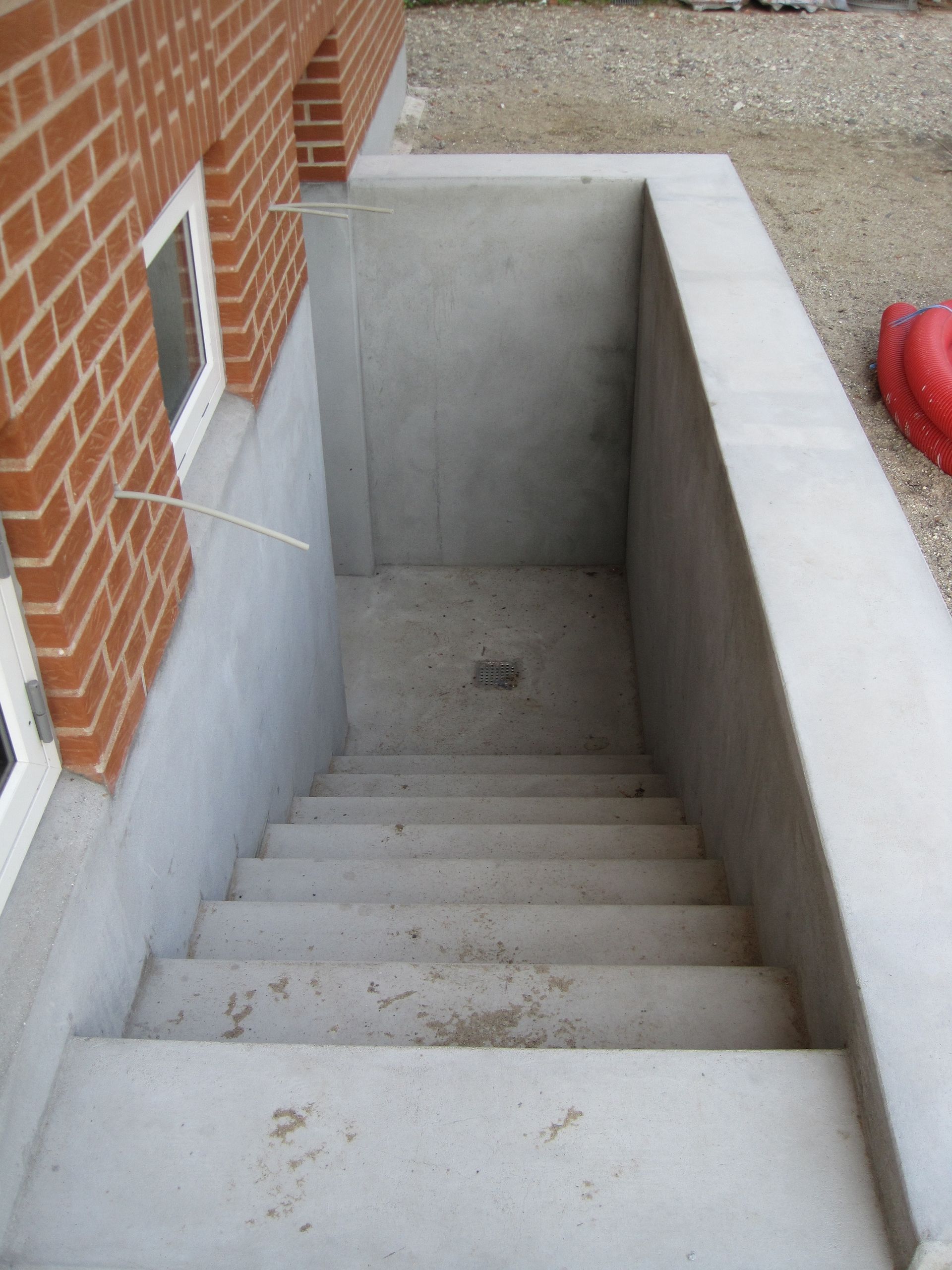 Betonkældertrappe, der fører ned, med murstensvæg og vindue i siden.