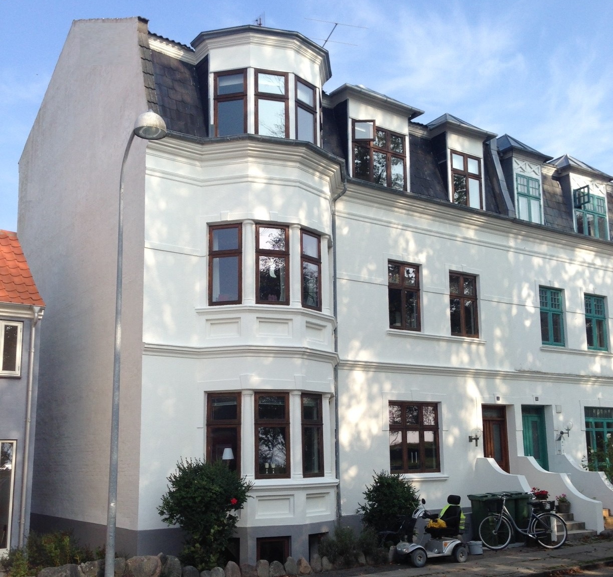 Hvidt rækkehus med karnappvinduer, mørkt tag og flere etager på en solskinsdag.