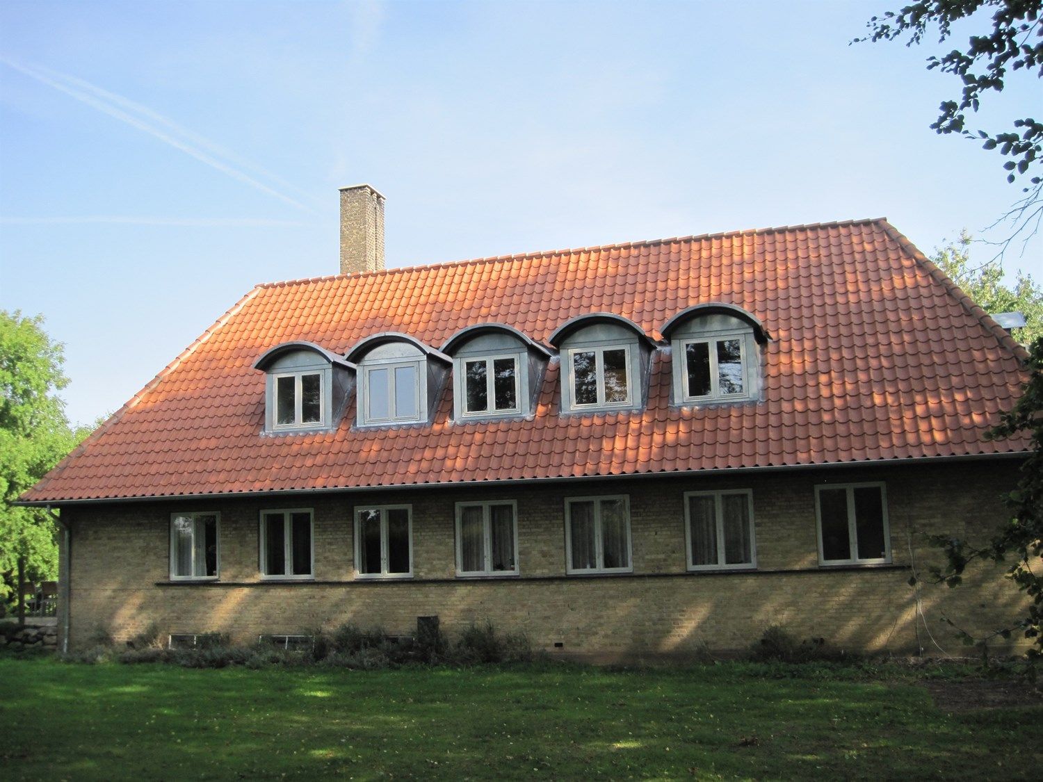 Brun murstensbygning med rødt tegltag, kvistvinduer og skorsten. Grøn græsplæne og træer.