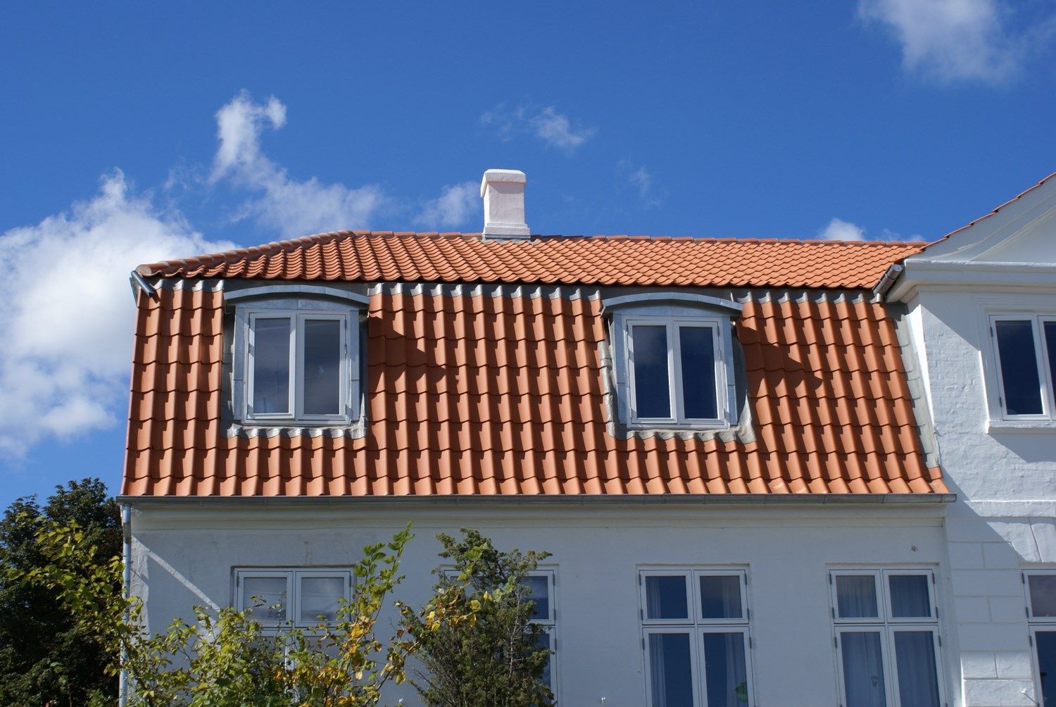 Hvid bygning med orange tegltag, to kvistvinduer og blå himmel.