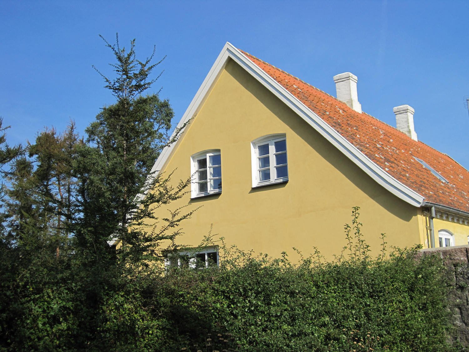 Gult hus med hvide lister, rødt tegltag, to skorstene og grønt løv mod en blå himmel.