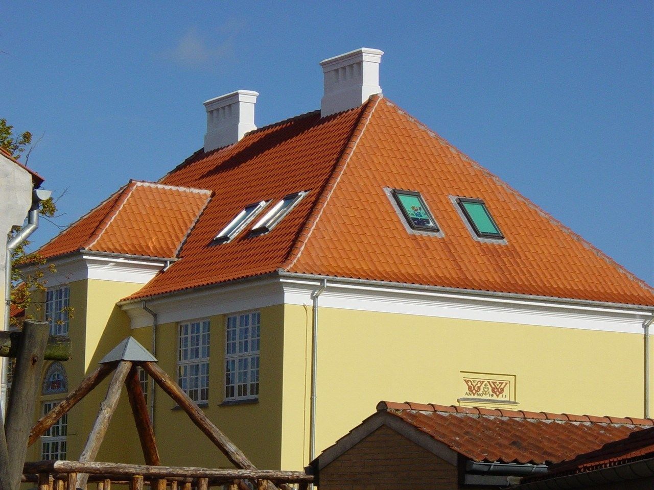 Gul bygning med orange tegltag, hvide lister, to skorstene og ovenlysvinduer mod en blå himmel.