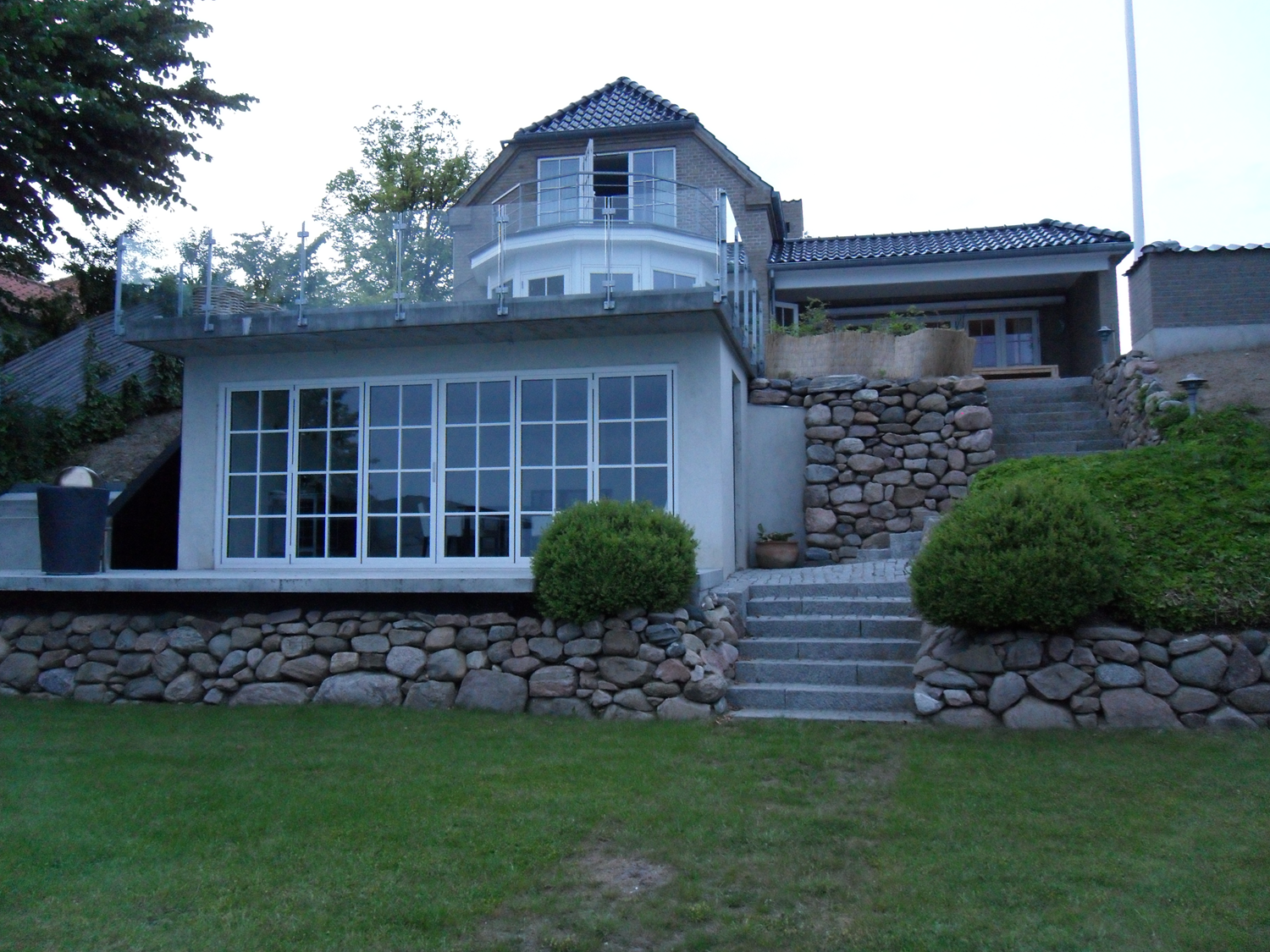 Hus med flere etager, stenmure og et solrum med glaspaneler. Grøn græsplæne i forgrunden og træer i baggrunden.