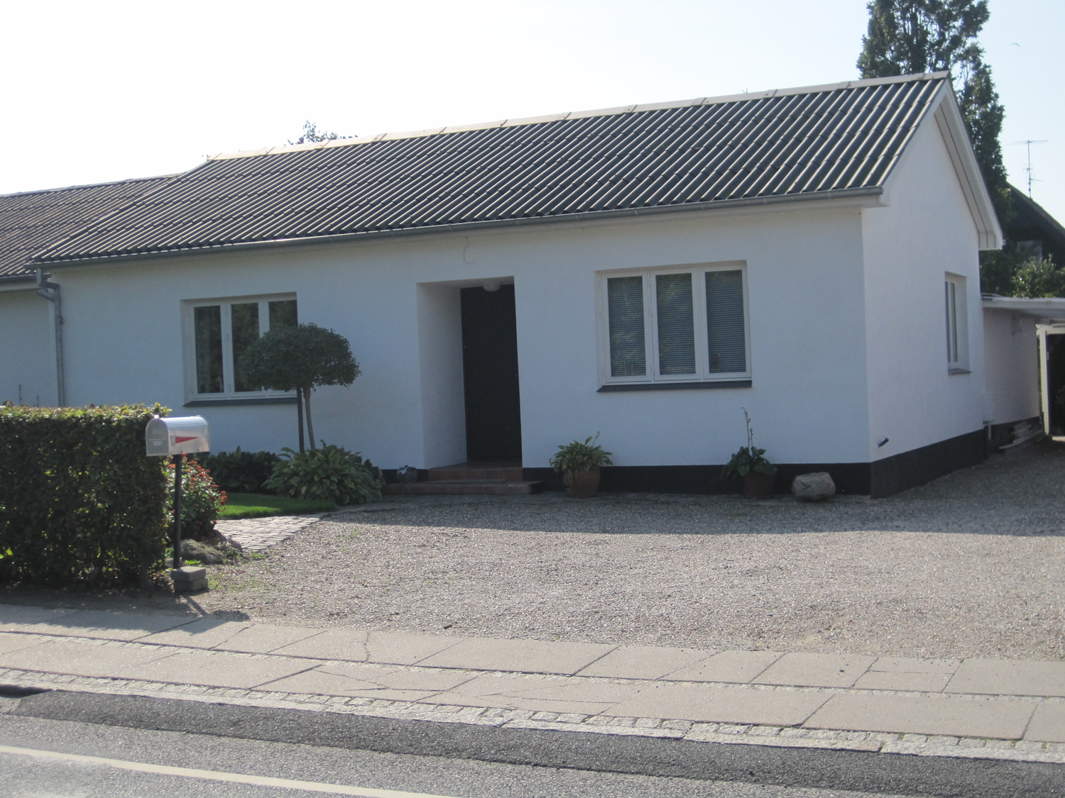 Hvid bungalow med mørkt tag, grå grusindkørsel, lille træ, postkasse og kort hæk.
