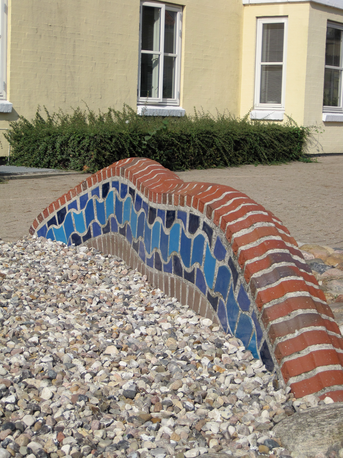 En buet have af mursten og blå fliser ligger foran en gul bygning med hvide vinduer i grus.