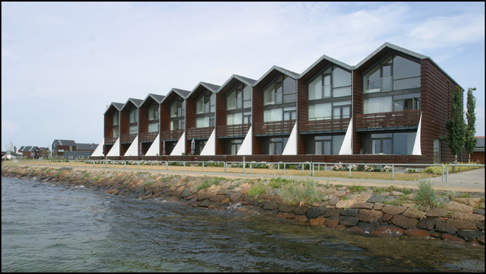 Række af moderne brune huse med spidse tage ved en havnefront.