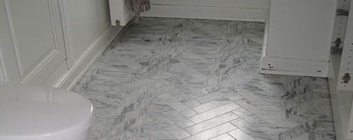 Gråt og hvidt marmorgulv i et badeværelse. Hvid væg med fodliste og et synligt toilet.