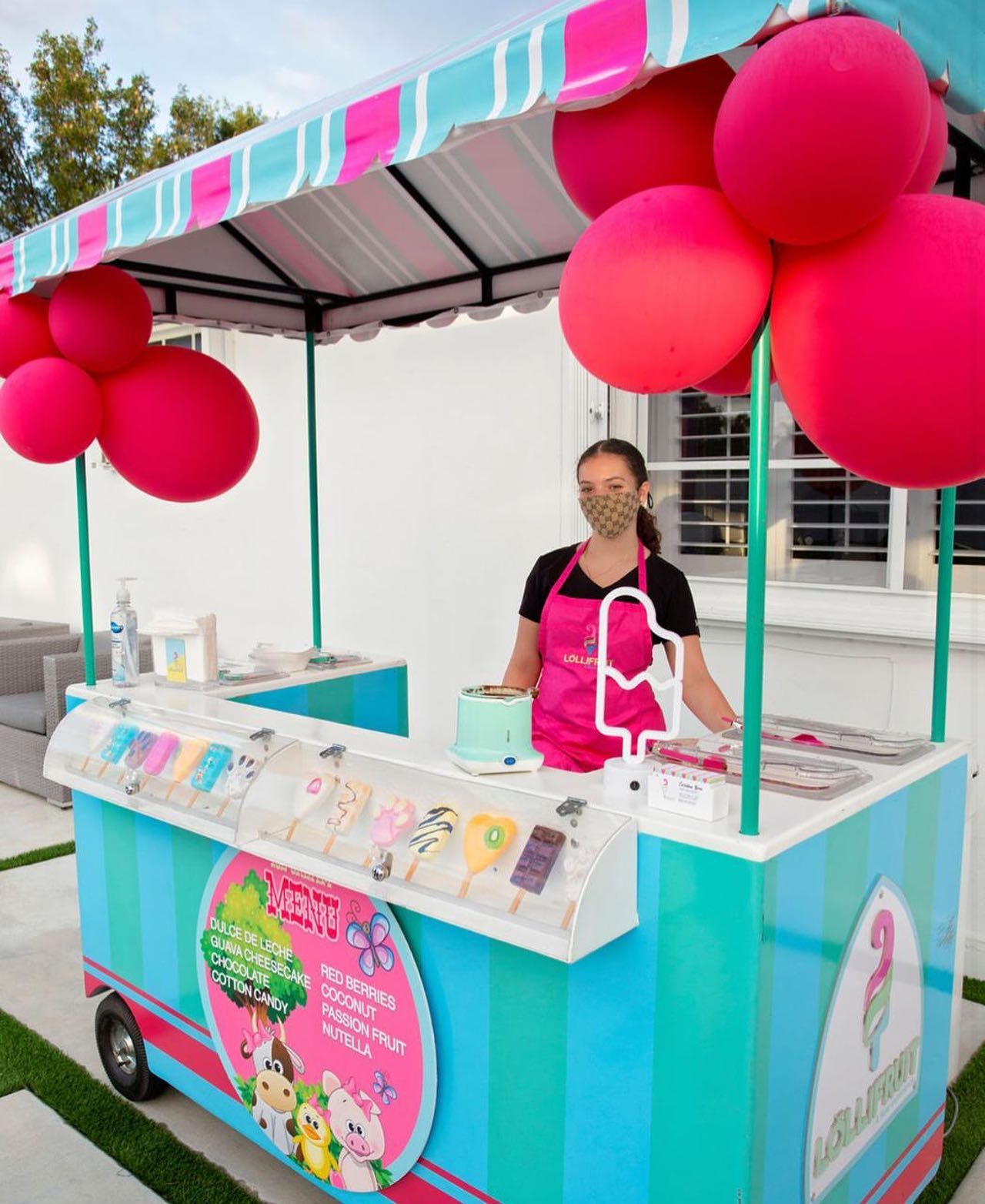 A woman is standing behind an ice cream cart with pink balloons.
