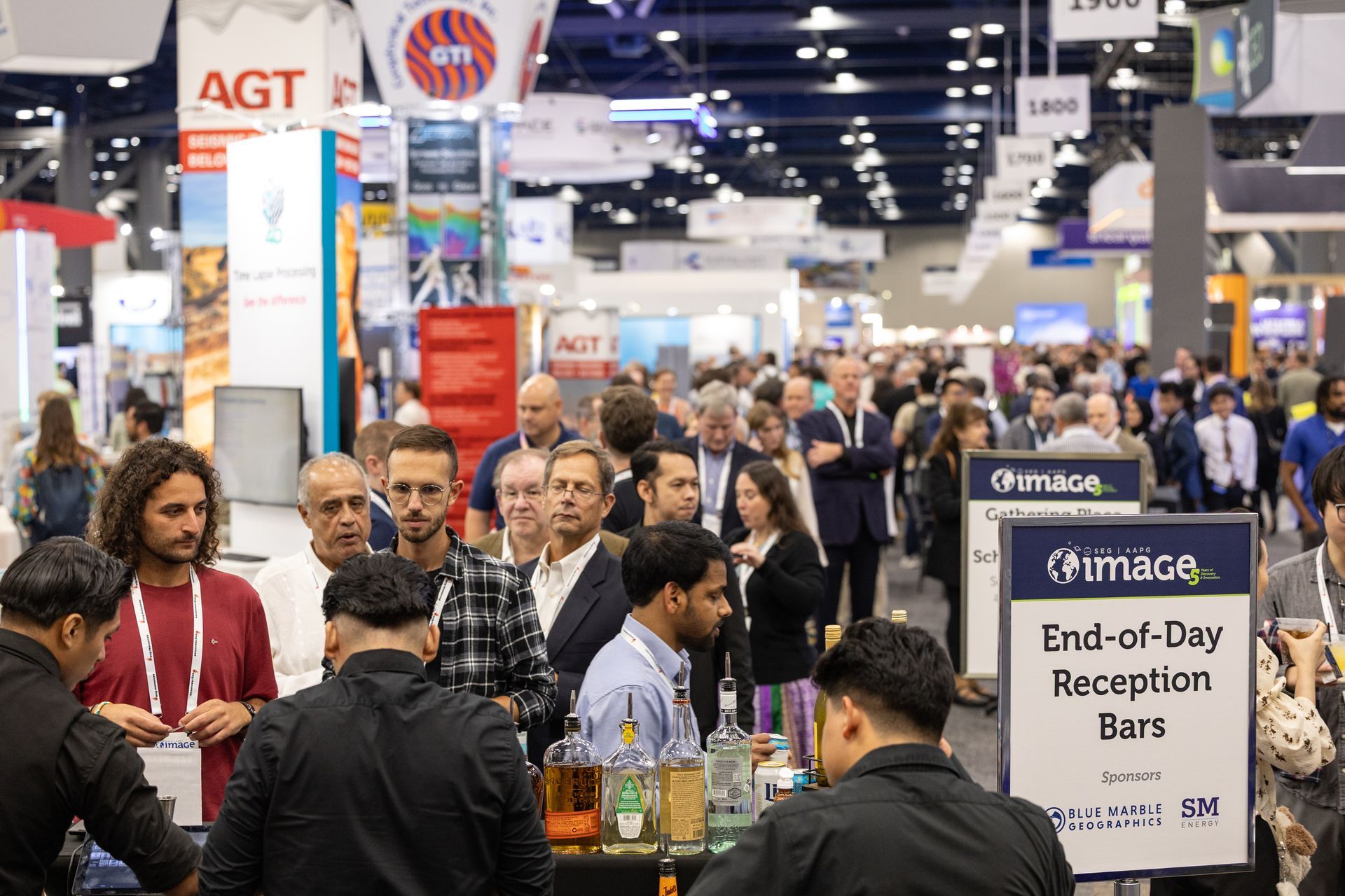 People at a crowded convention; bar with drinks in the foreground. Signs and booths in background.