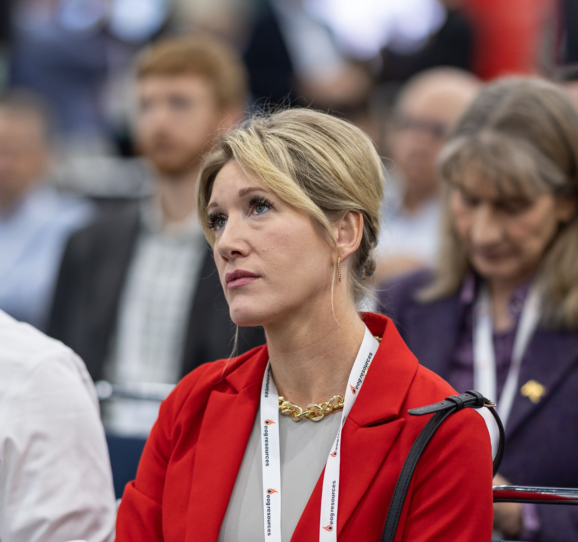 Woman in red blazer, gold necklace, looking off-camera at a conference.
