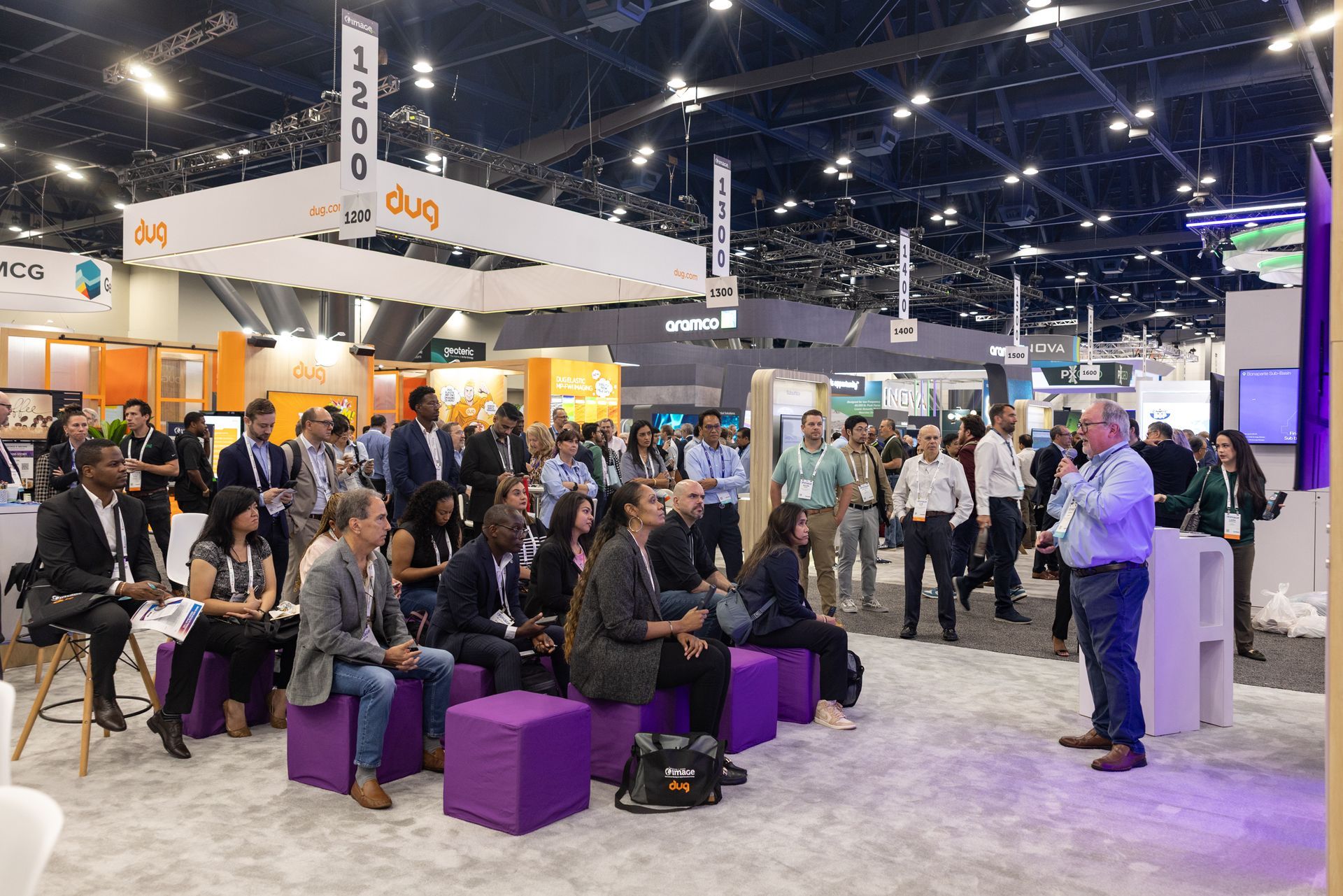 A speaker addresses a crowd at a trade show. People listen while seated and standing near booths.