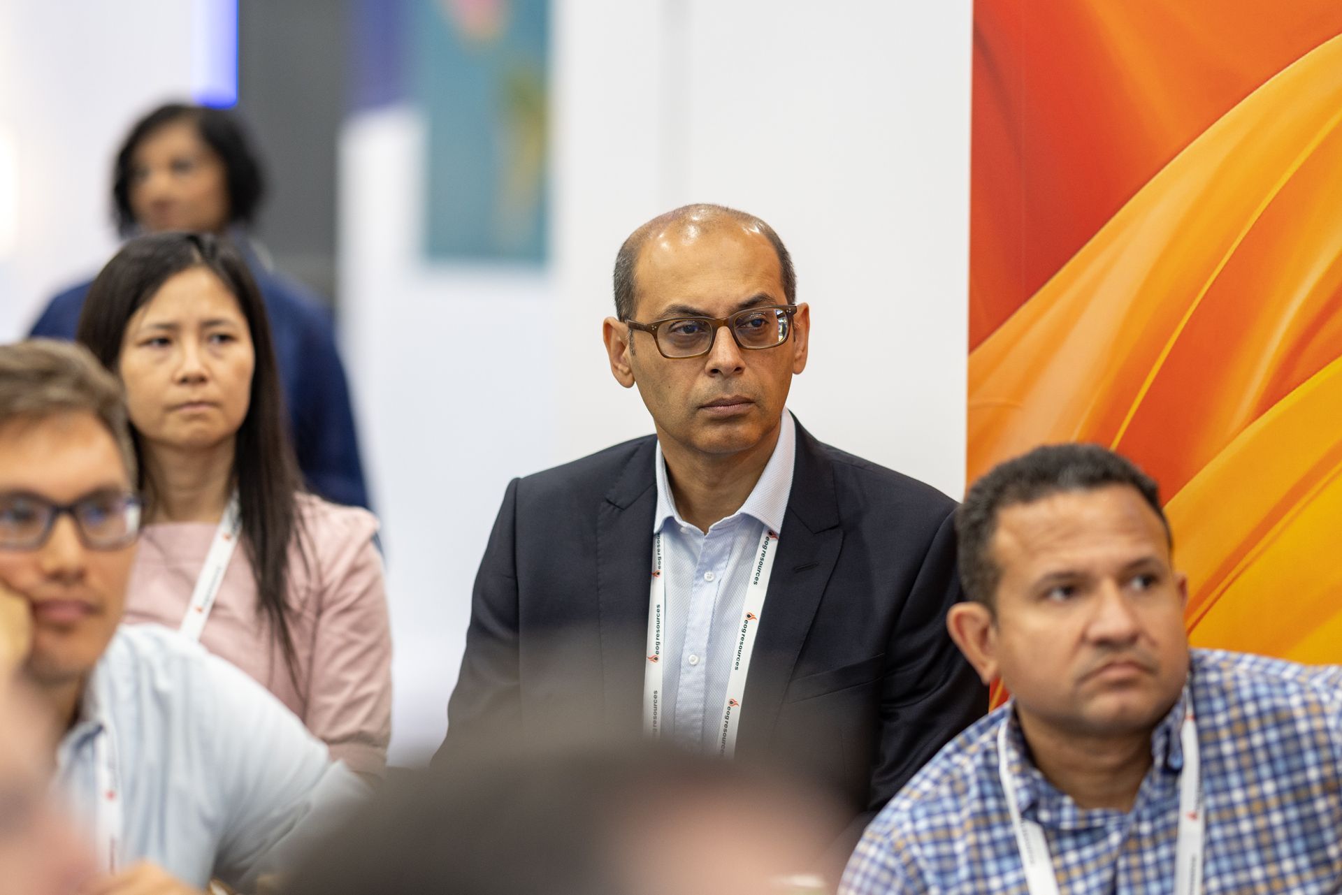 Attendees at a conference. Man in suit looking forward, others listen intently. Bright orange background.