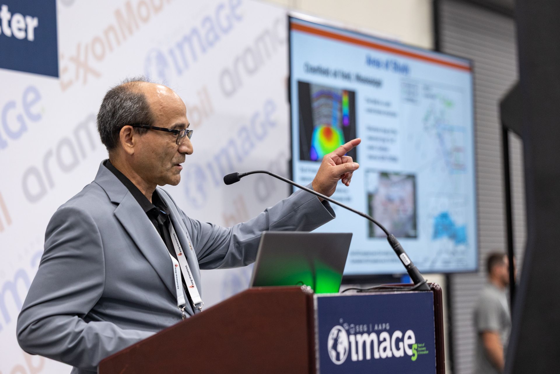 Man giving a presentation at a conference, pointing at a screen. Podium, laptop, and logo visible.