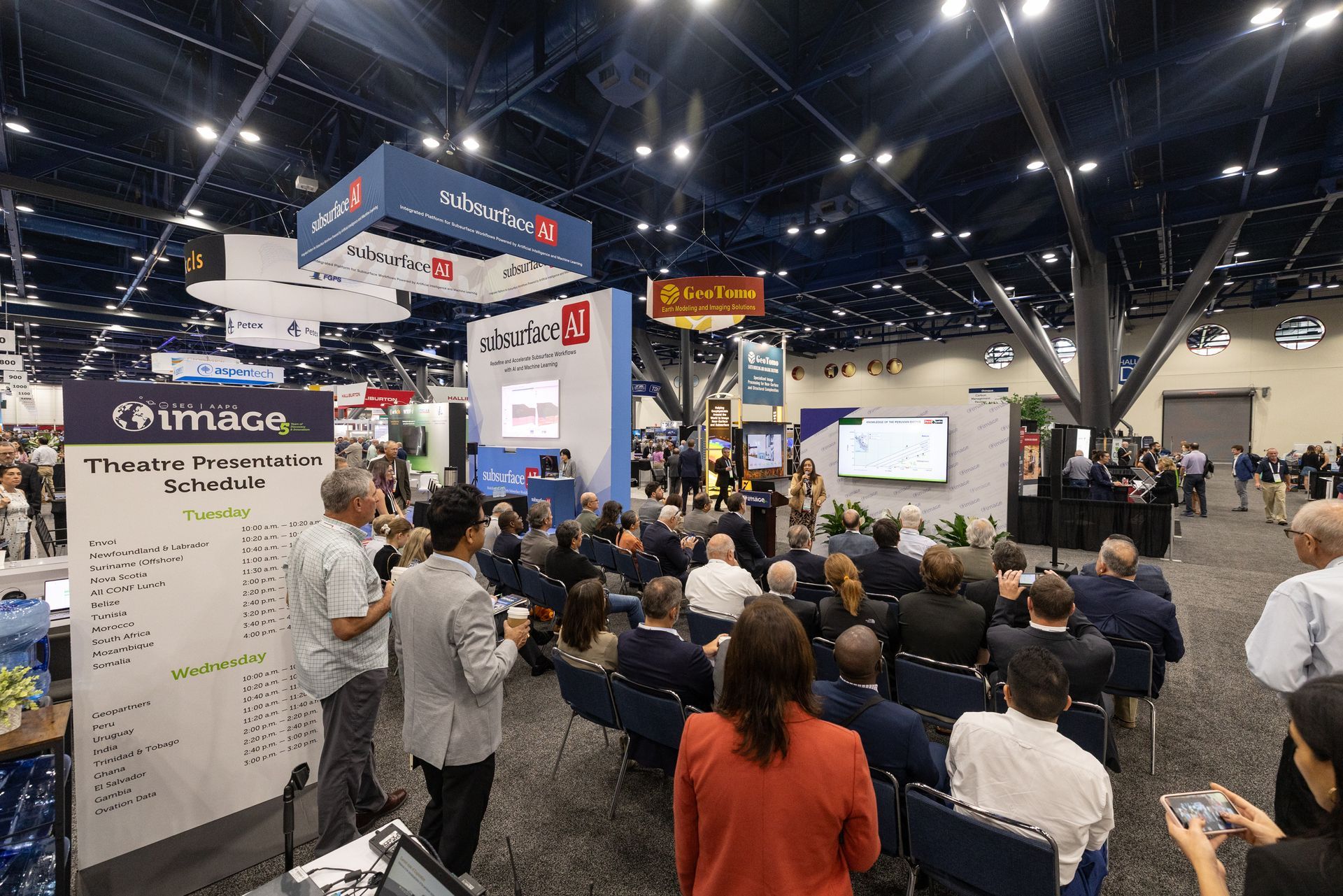 Large convention hall with a seated audience, a speaker at a podium, and company booths visible.
