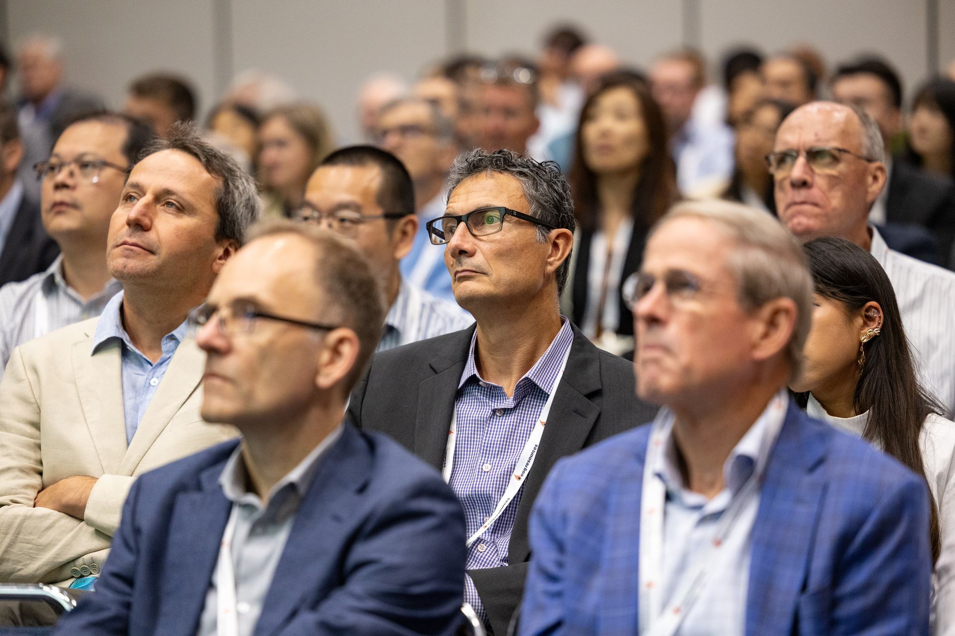Audience members attentively listen at a conference; some wear glasses and business attire.