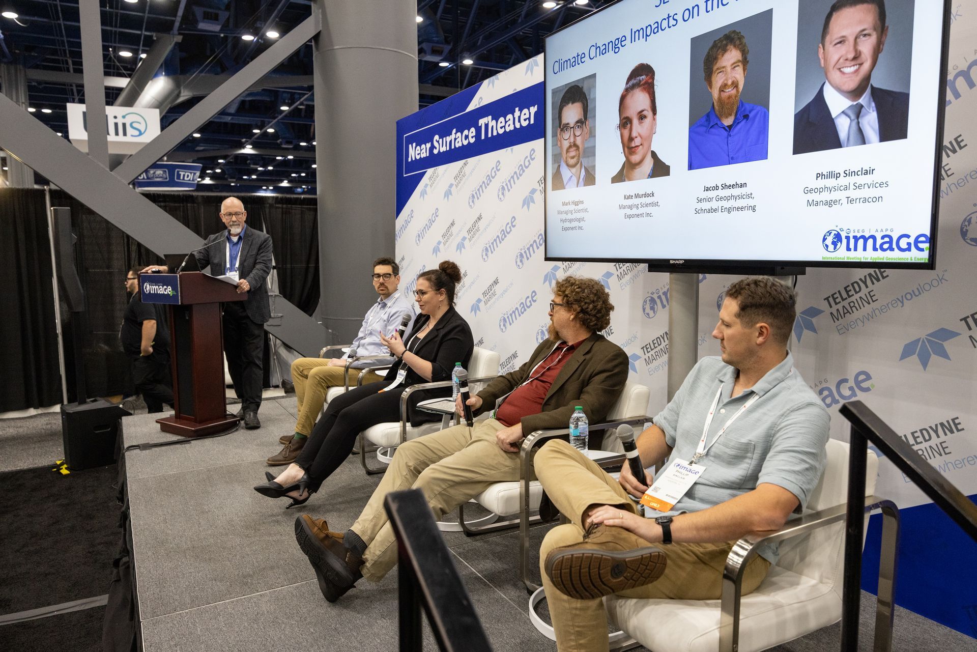 Panel discussion at a convention: speaker at a podium, panelists seated. Screen shows headshots.