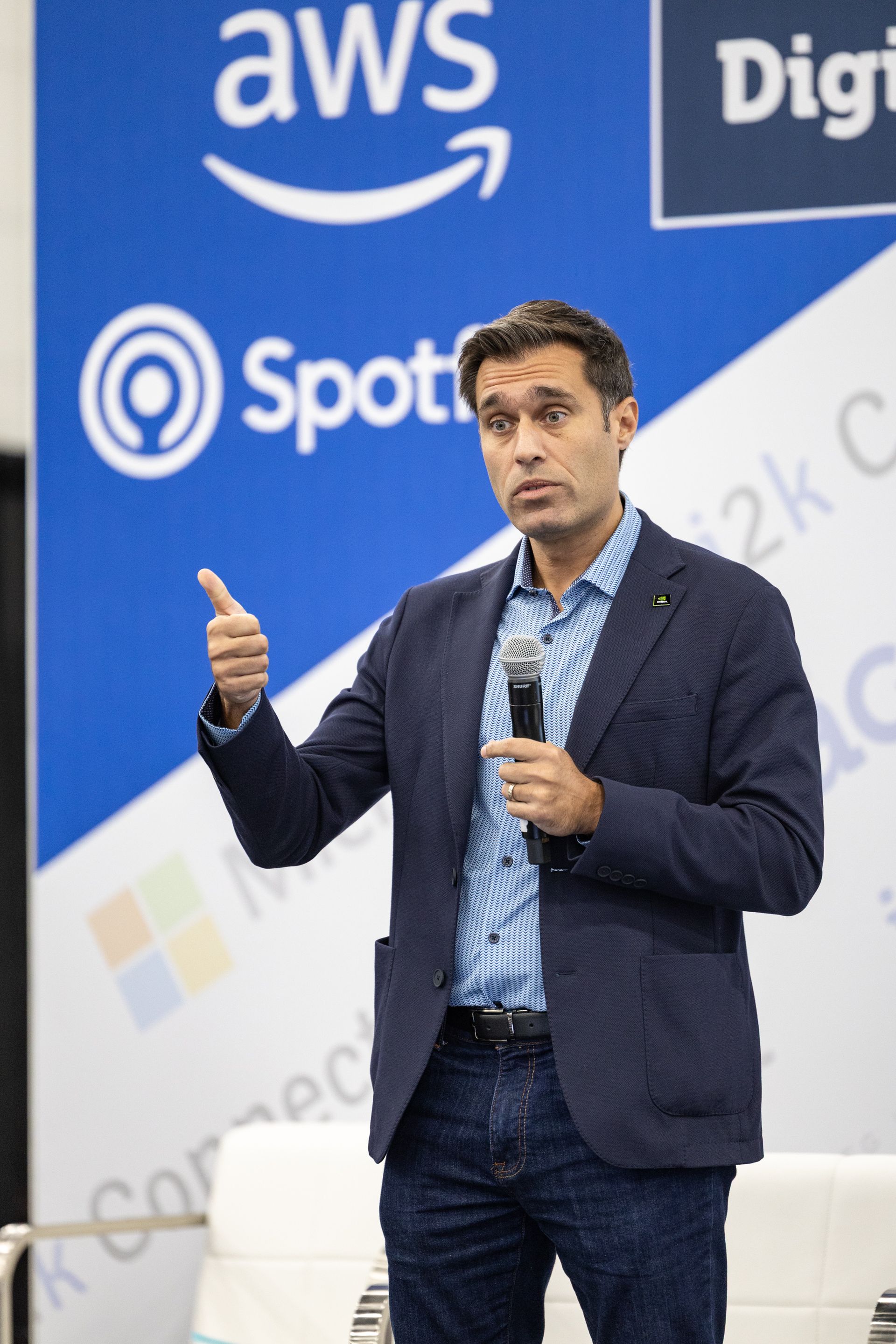Man in blazer speaking into a microphone at a conference with company logos in the background.