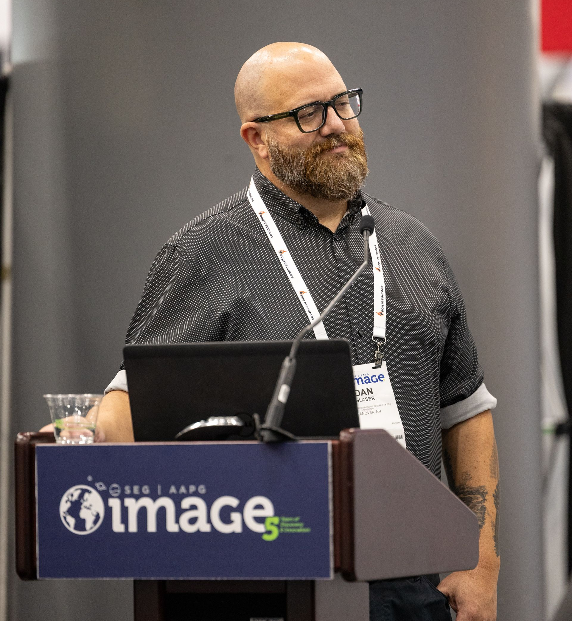 Man with beard and glasses speaking at a podium with a laptop, the logo