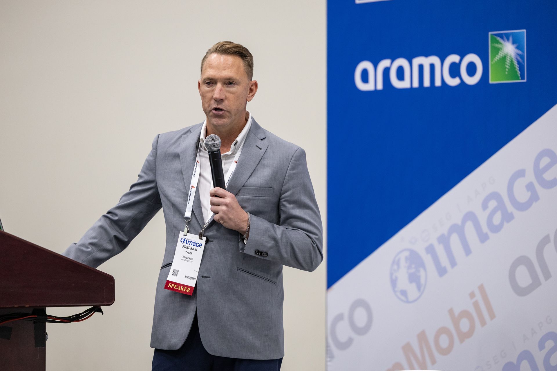 Man speaking at a podium, Aramco banner in background. He wears a gray blazer, holding a microphone.