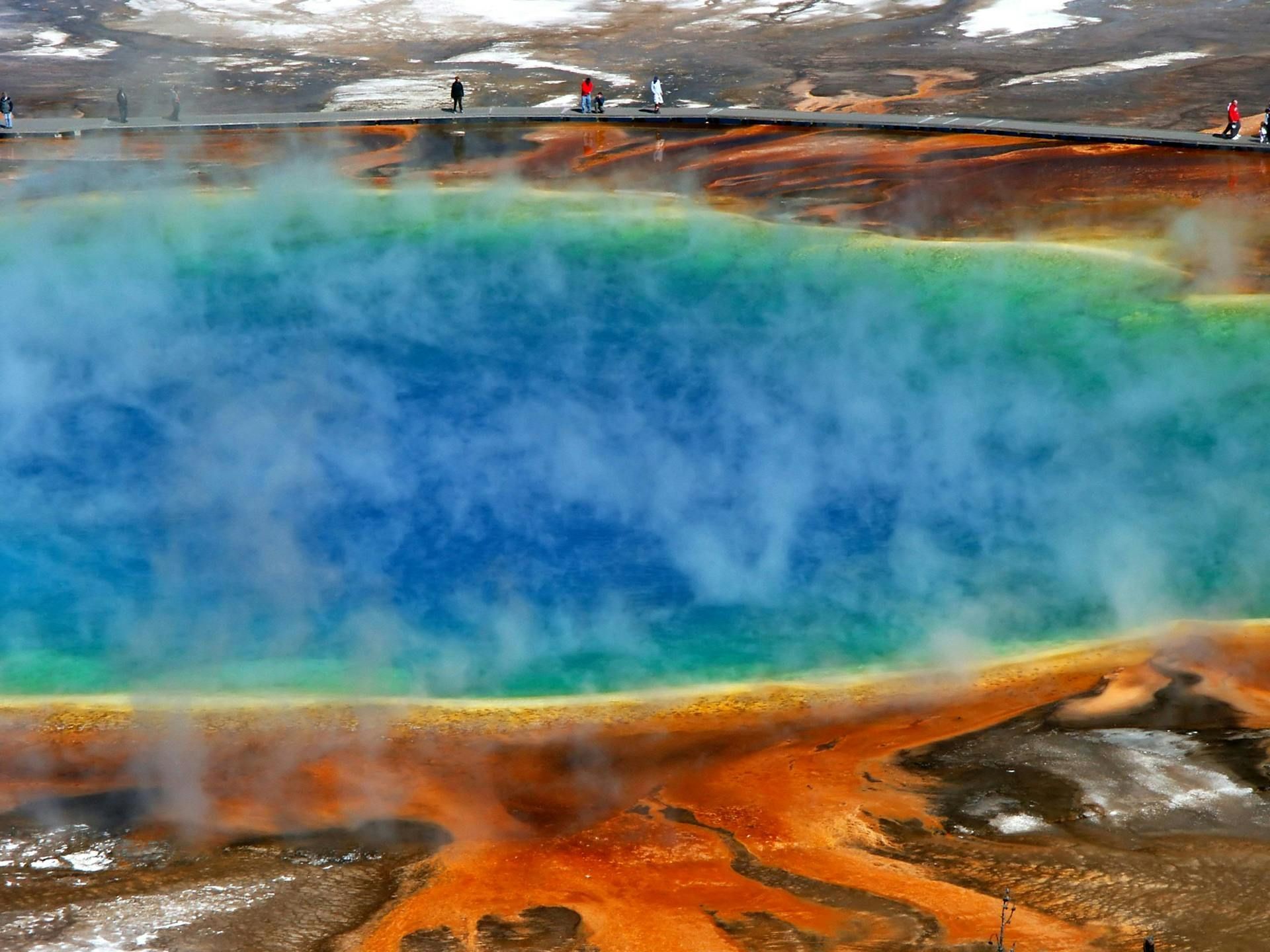 A colorful geothermal pool at Yellowstone National Park, with vibrant blue water and orange/brown mineral deposits.