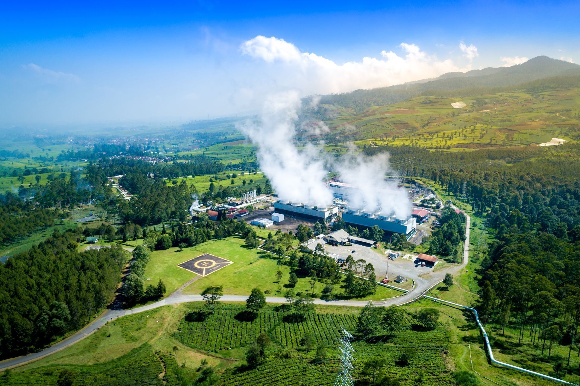 Geothermal power plant in a green landscape with steam rising, a helipad, and a road.