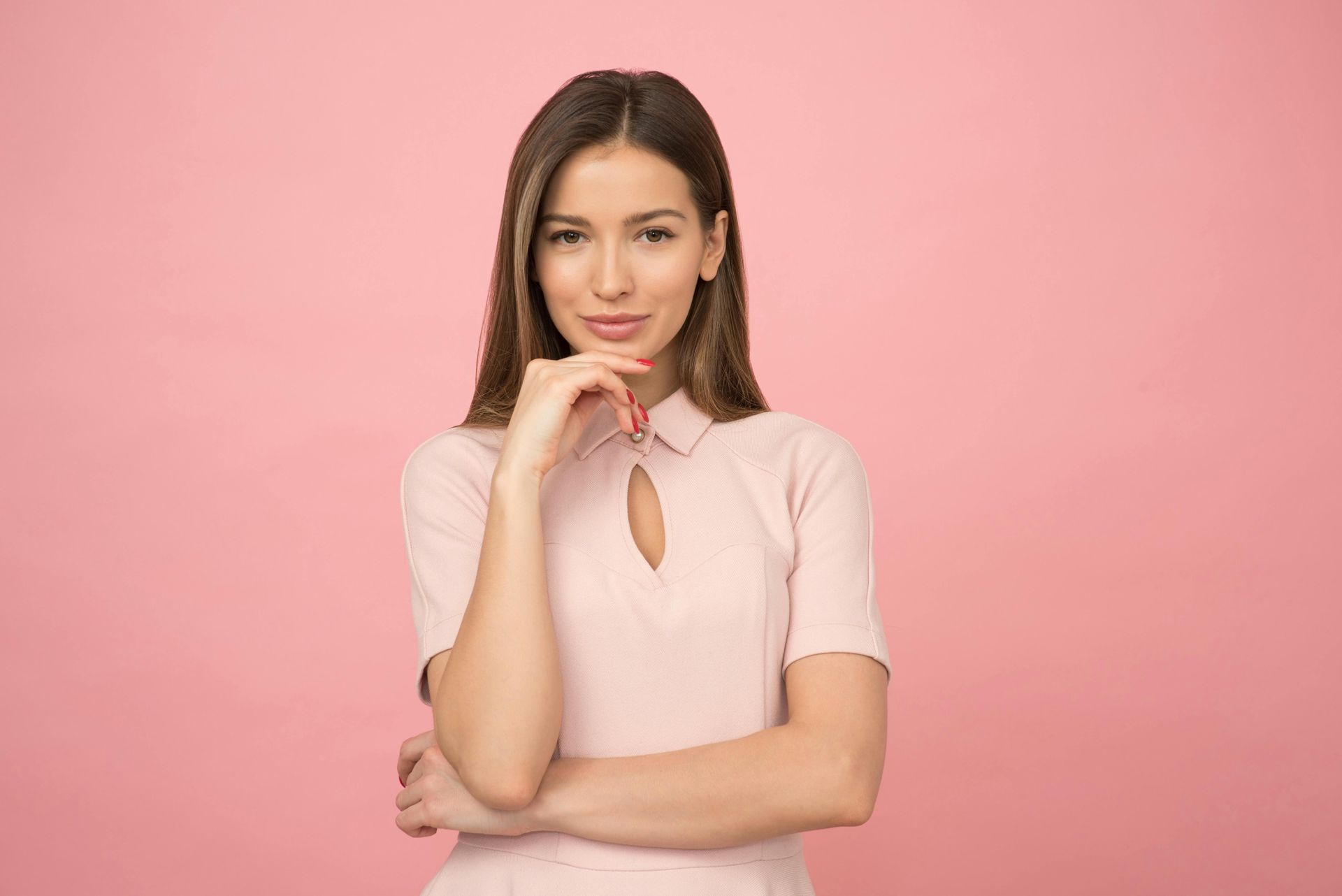 A woman in a pink shirt is standing with her arms crossed and her hand on her chin.