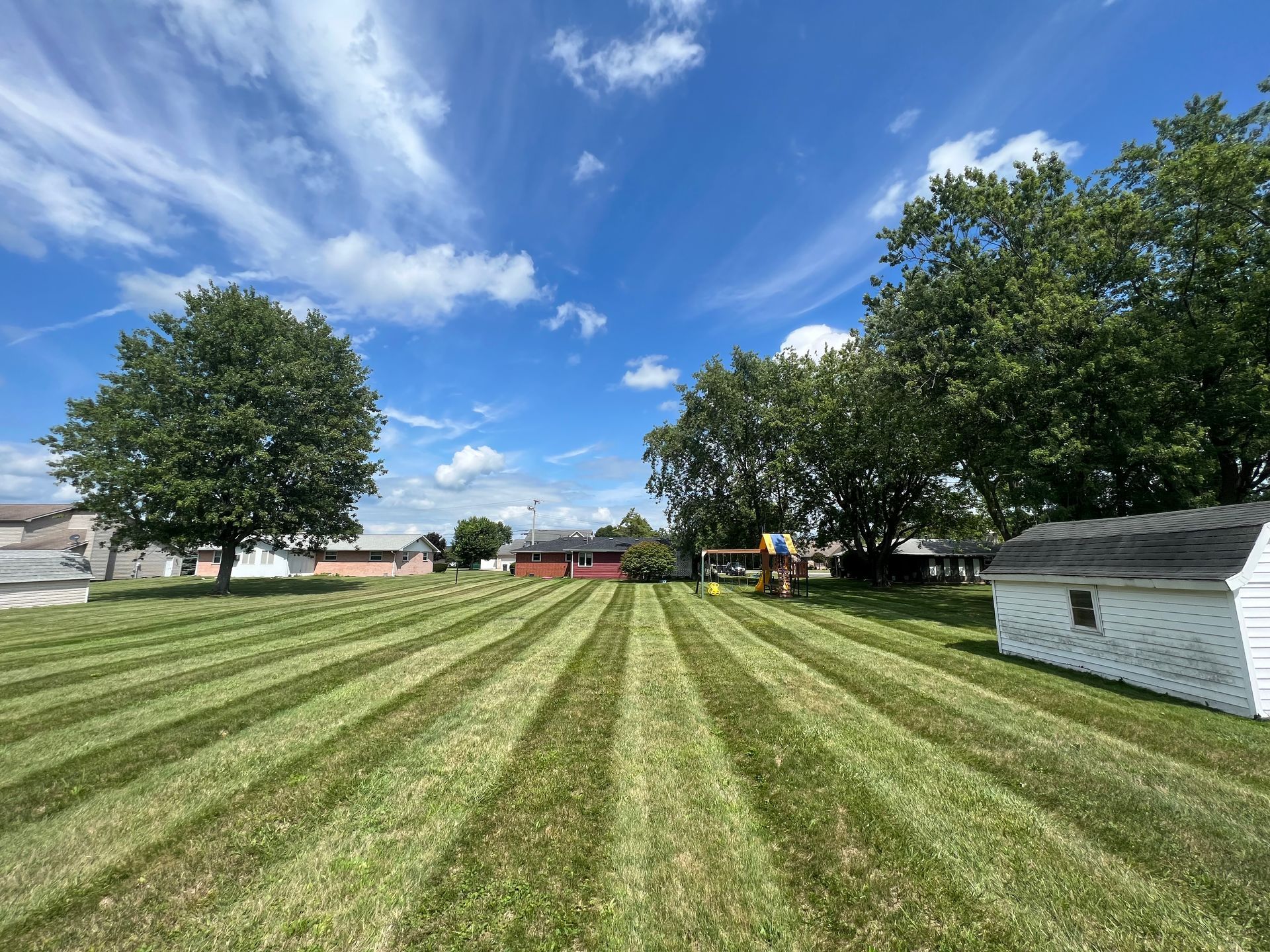 Lawn with freshly mown stripes, under a bright blue sky with fluffy clouds; trees and buildings in the background.