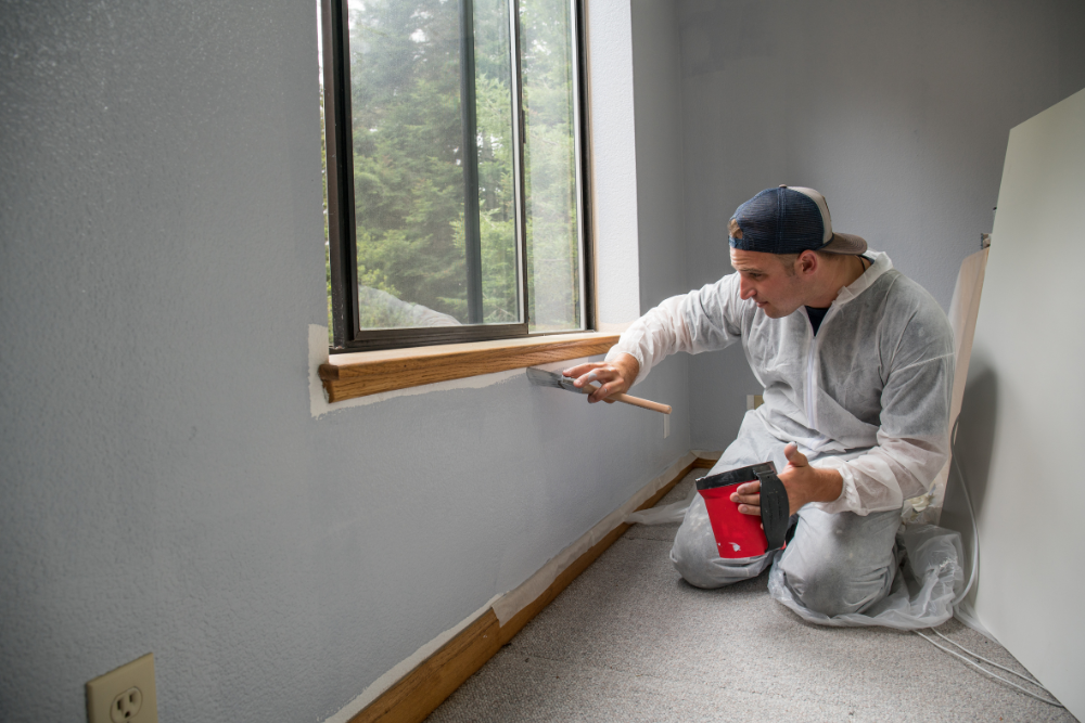 A man is kneeling down in a room painting a wall.