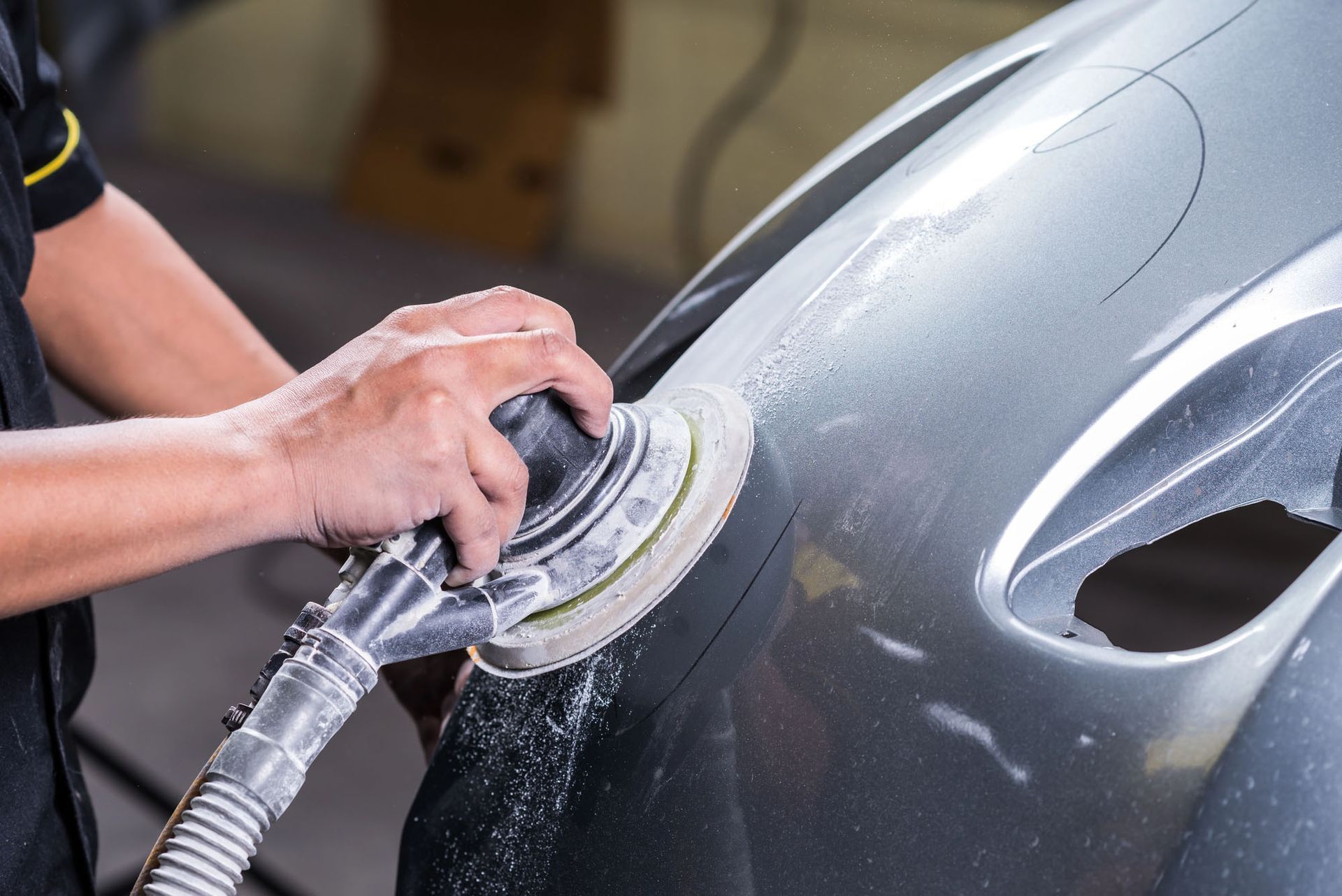 Una persona leviga un pannello grigio di un'auto con una levigatrice elettrica durante una riparazione della carrozzeria.