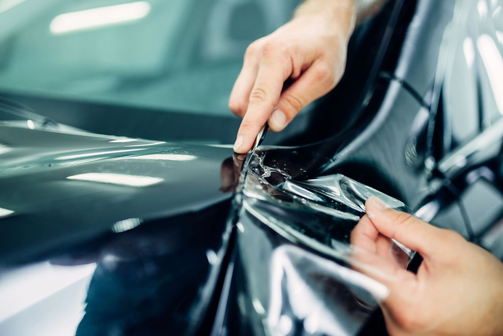 Mani che applicano una pellicola protettiva trasparente sul cofano scuro di un'auto.
