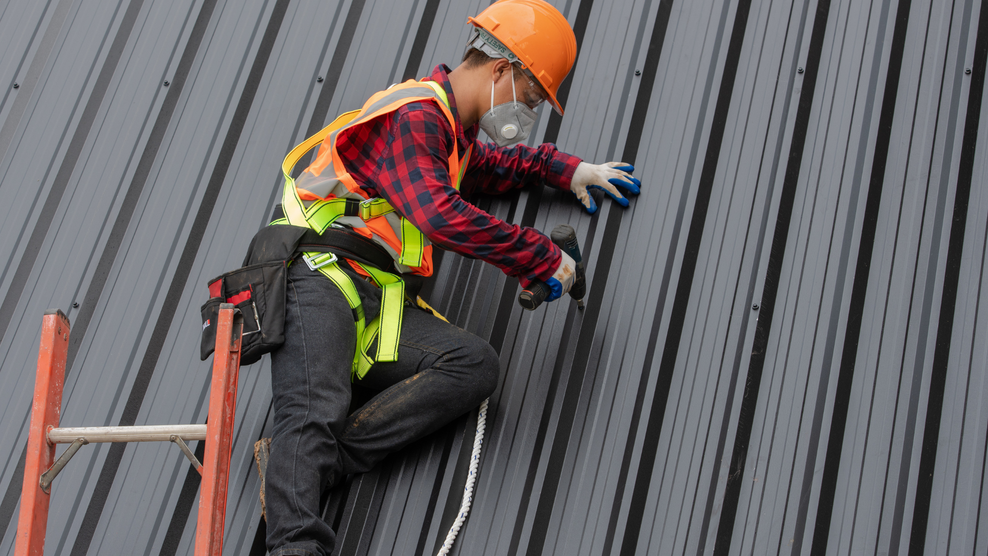 A worker wearing a hard hat, respirator, and safety harness secures metal wall panels while standing on a ladder.